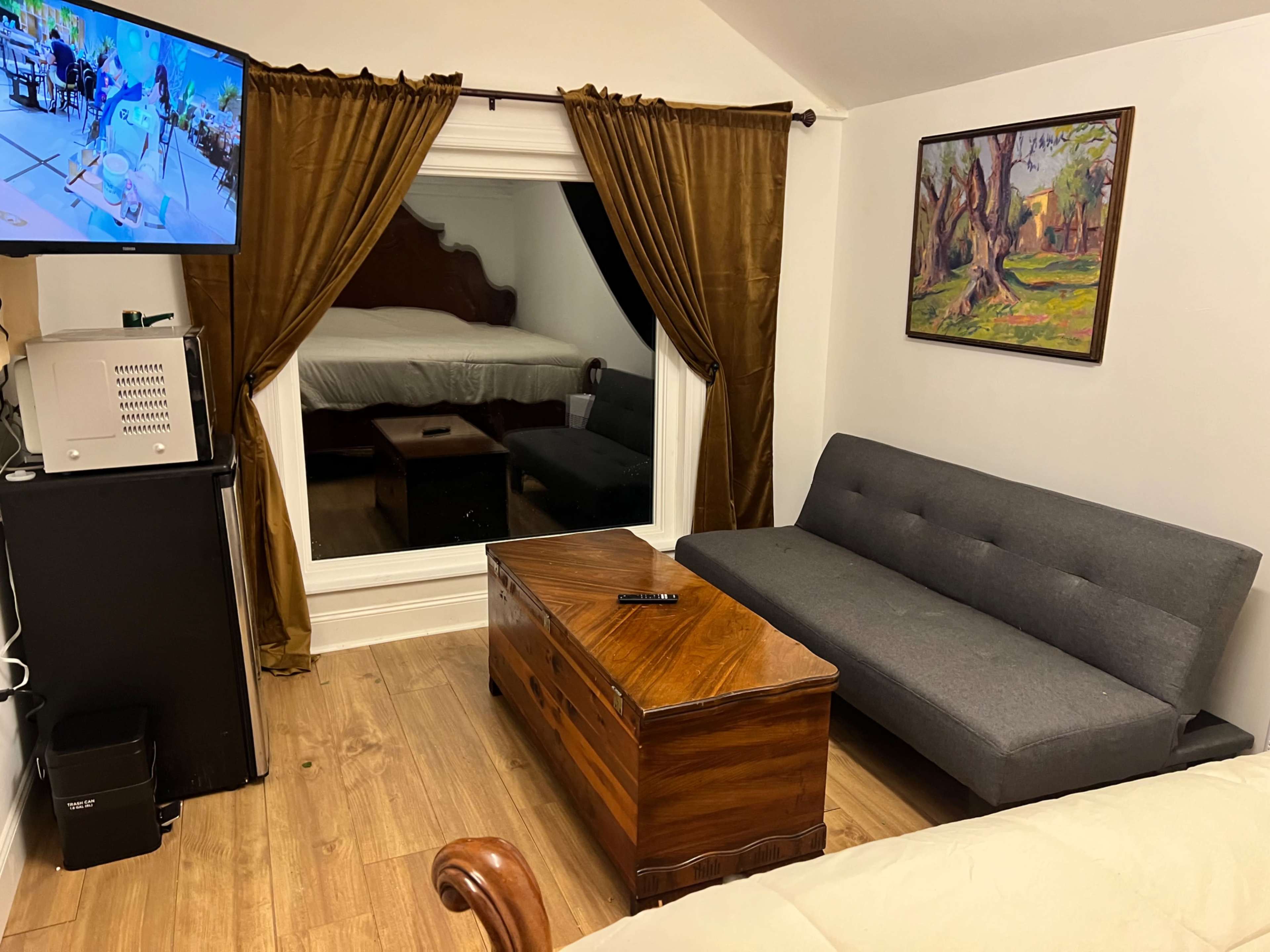 A modest room features a gray sofa, a wooden coffee table, and a television mounted on the wall, with a bed visible through a window.