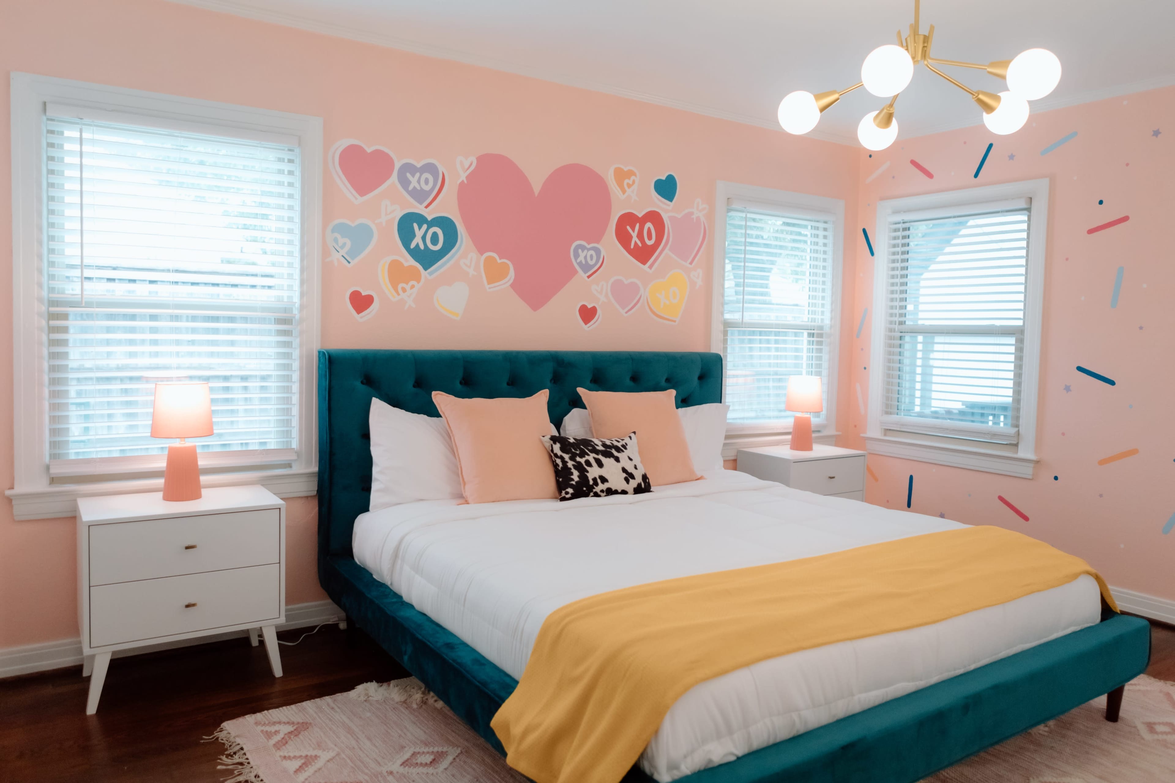The bedroom features a teal upholstered bed with a yellow blanket, pink walls adorned with heart and sprinkle designs, and two windows flanked by bedside tables with lamps.