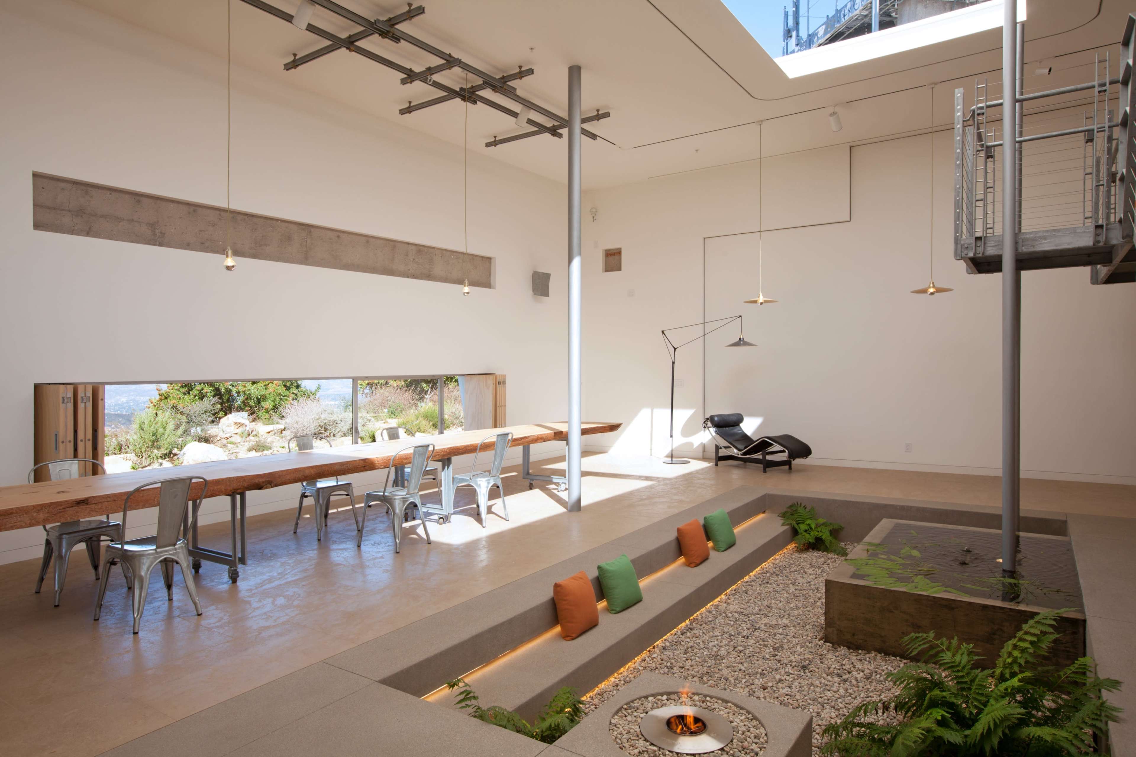 The image shows a spacious, modern interior with a long wooden dining table, metal chairs, and a central decorative area featuring a stone arrangement and a small fire pit.