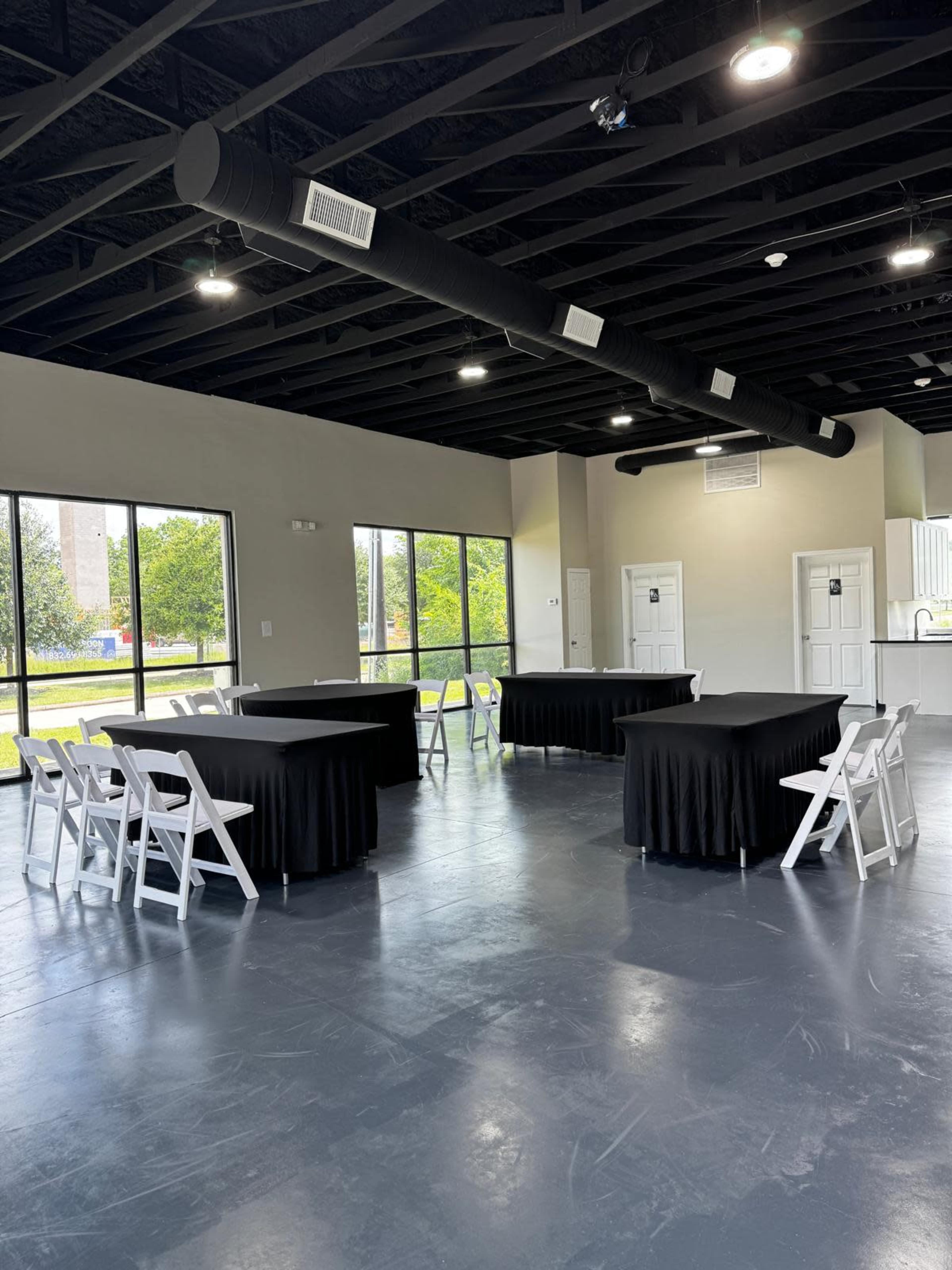 The image shows a spacious room with black tablecloths covering several tables and white folding chairs arranged around them, illuminated by overhead lights and large windows.