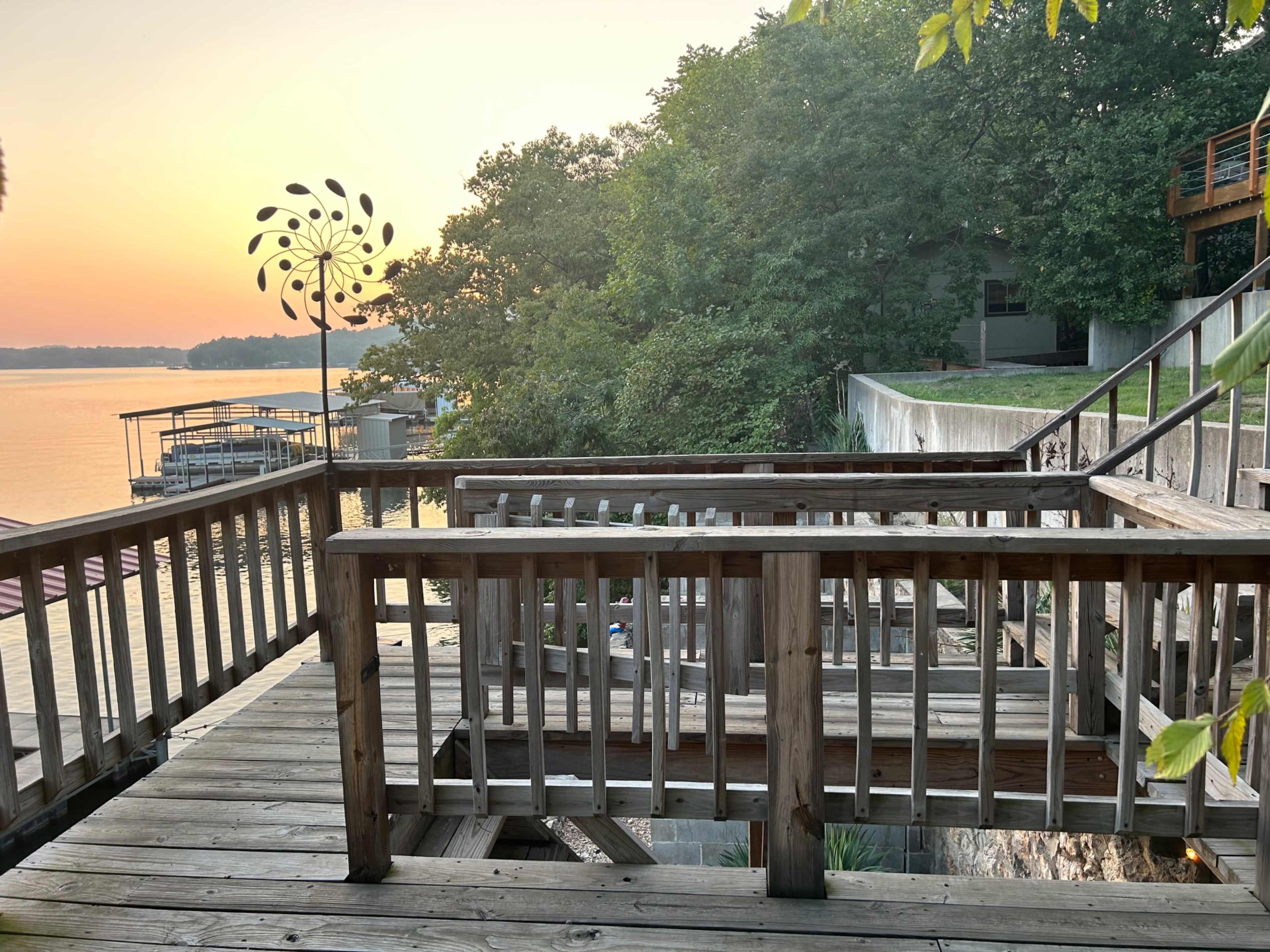 A wooden deck overlooks a calm lake at sunset, with trees lining the shoreline and a wind spinner in the background.