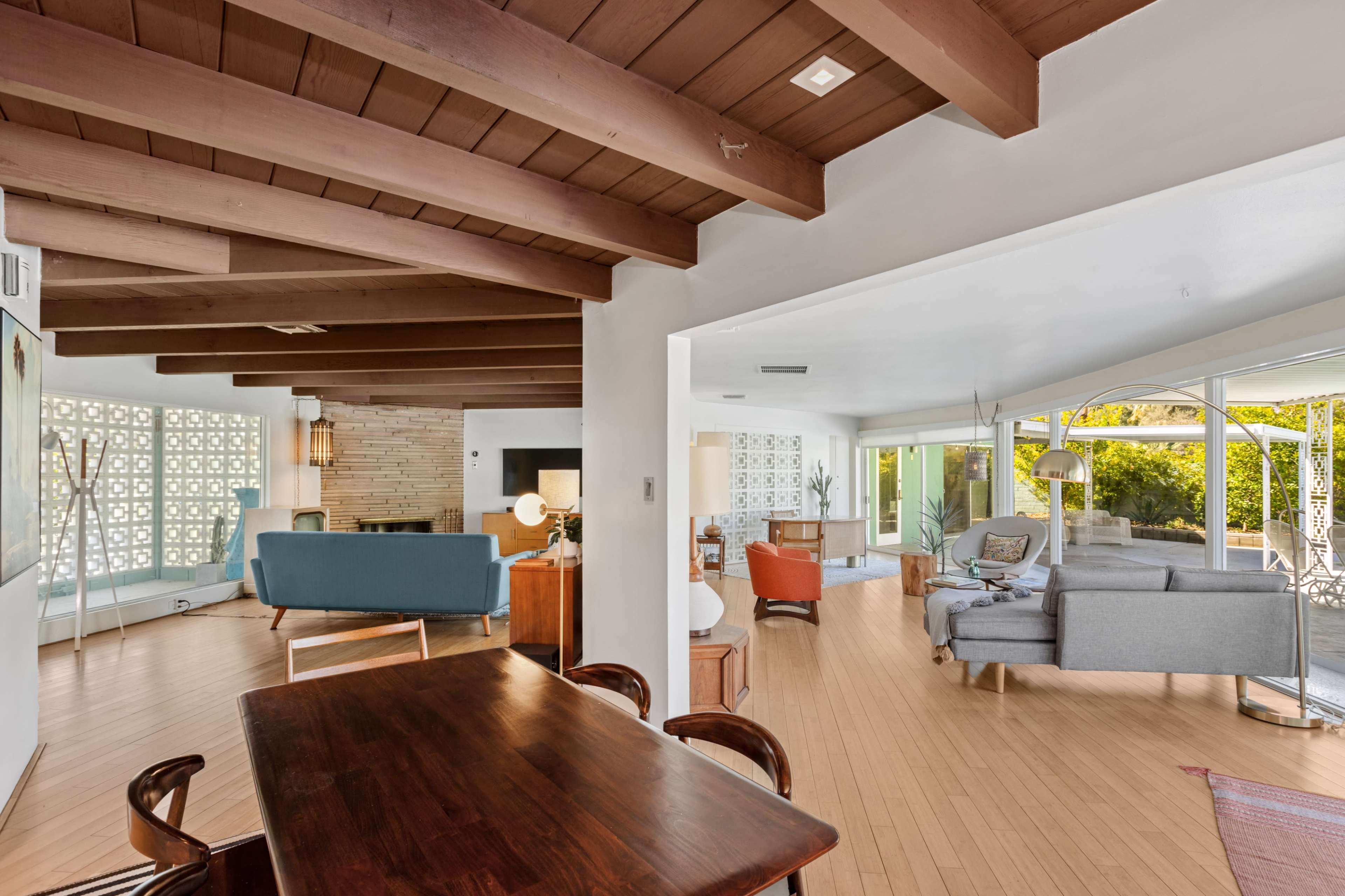 The image shows an open-concept living space featuring a dining area with wooden beams, a blue sofa, a fireplace, and large windows that lead to a patio.