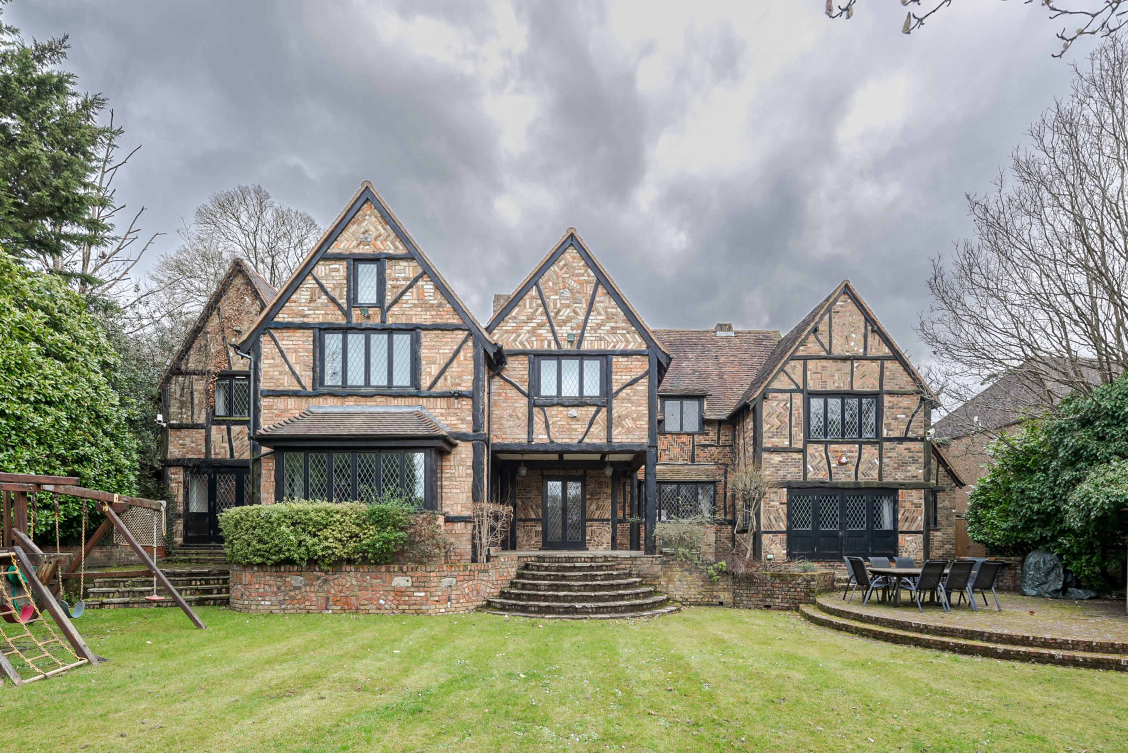 The image shows a large, traditional brick house with a steep, gabled roof and a spacious lawn in front, surrounded by trees and a patio area with outdoor furniture.