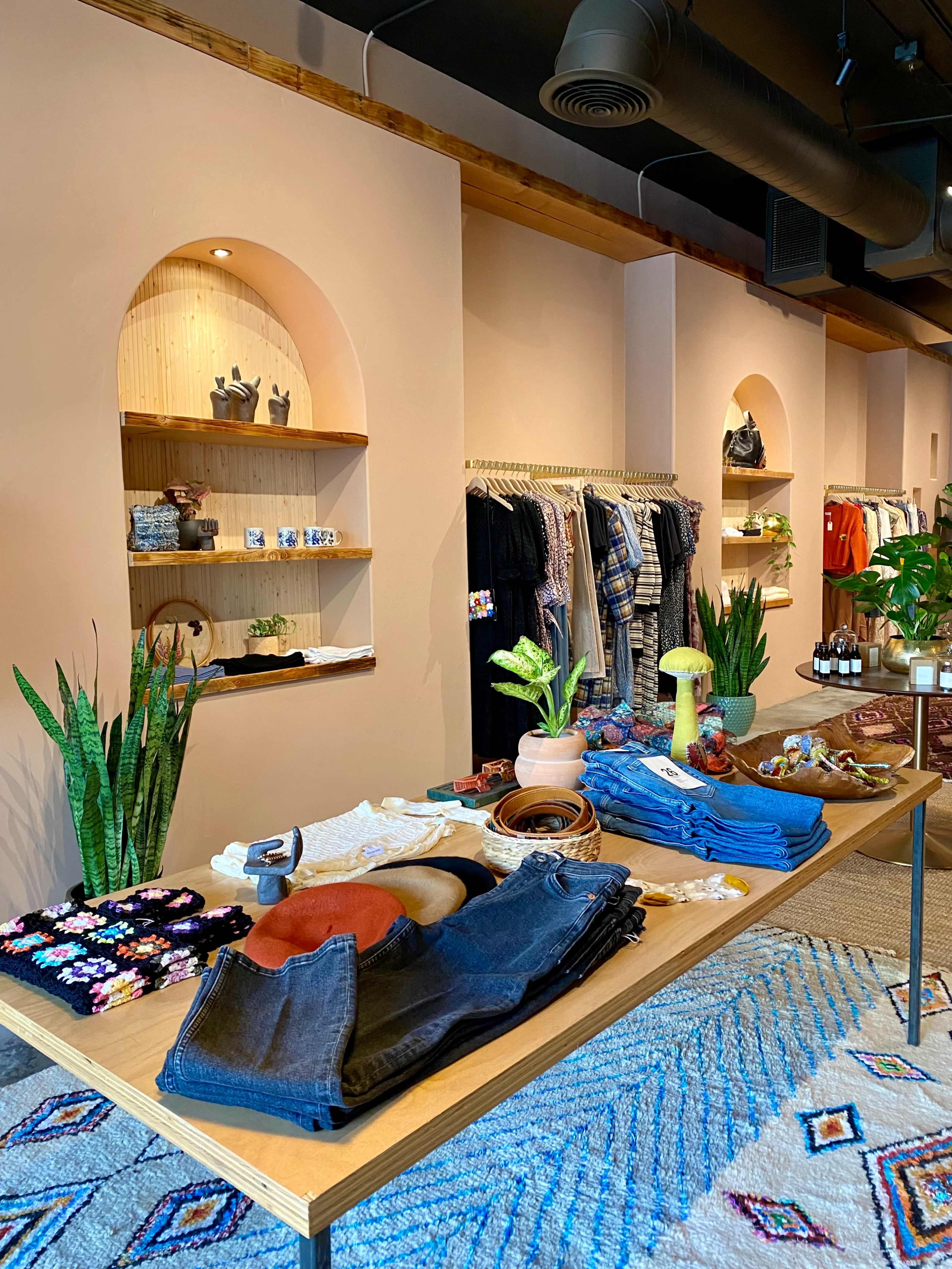 The image shows a retail display with neatly arranged clothing and accessories on a wooden table, set against a backdrop of arched shelves filled with decorative items.
