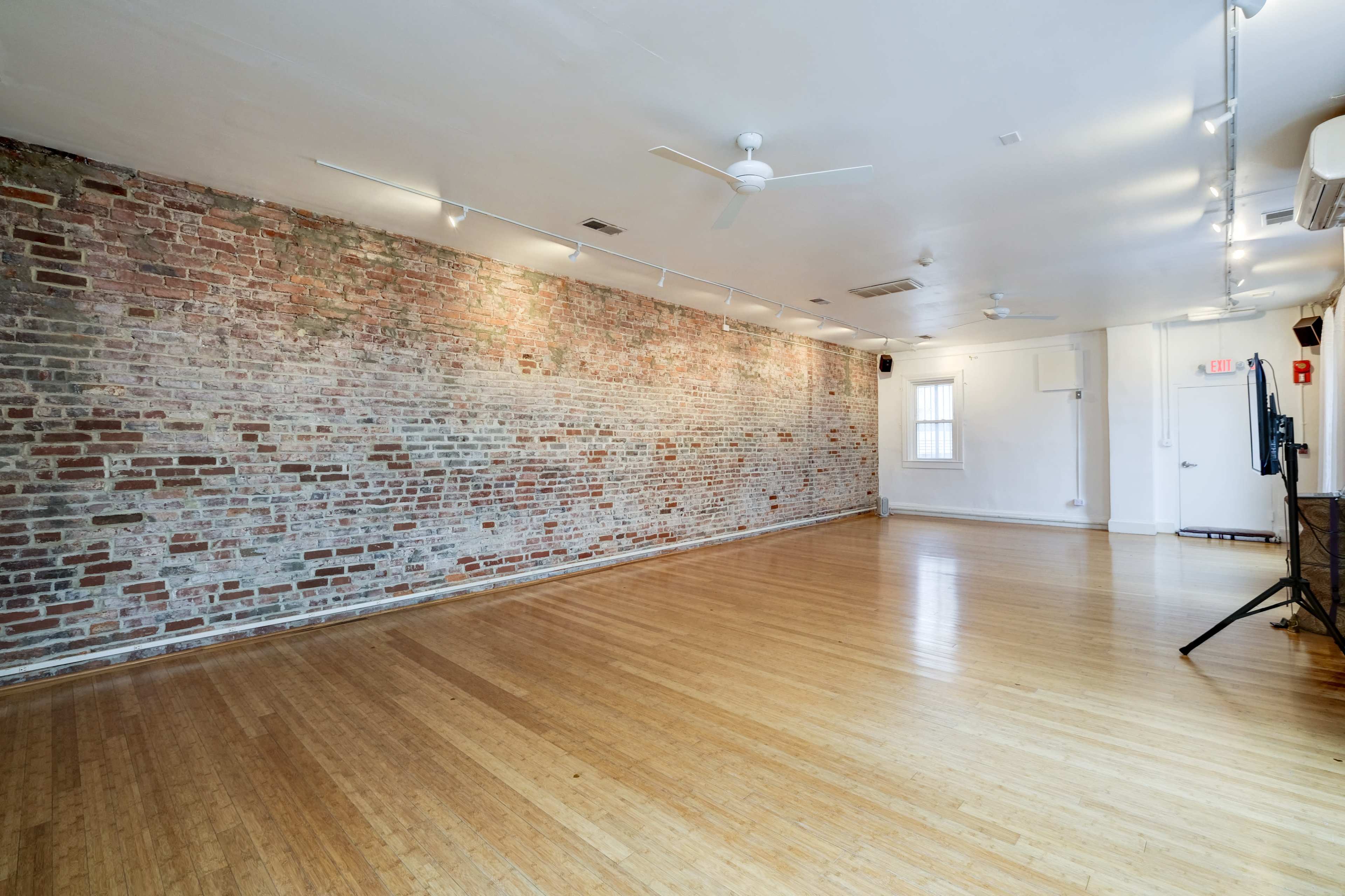An empty room with hardwood flooring and a brick wall, illuminated by ceiling lights.