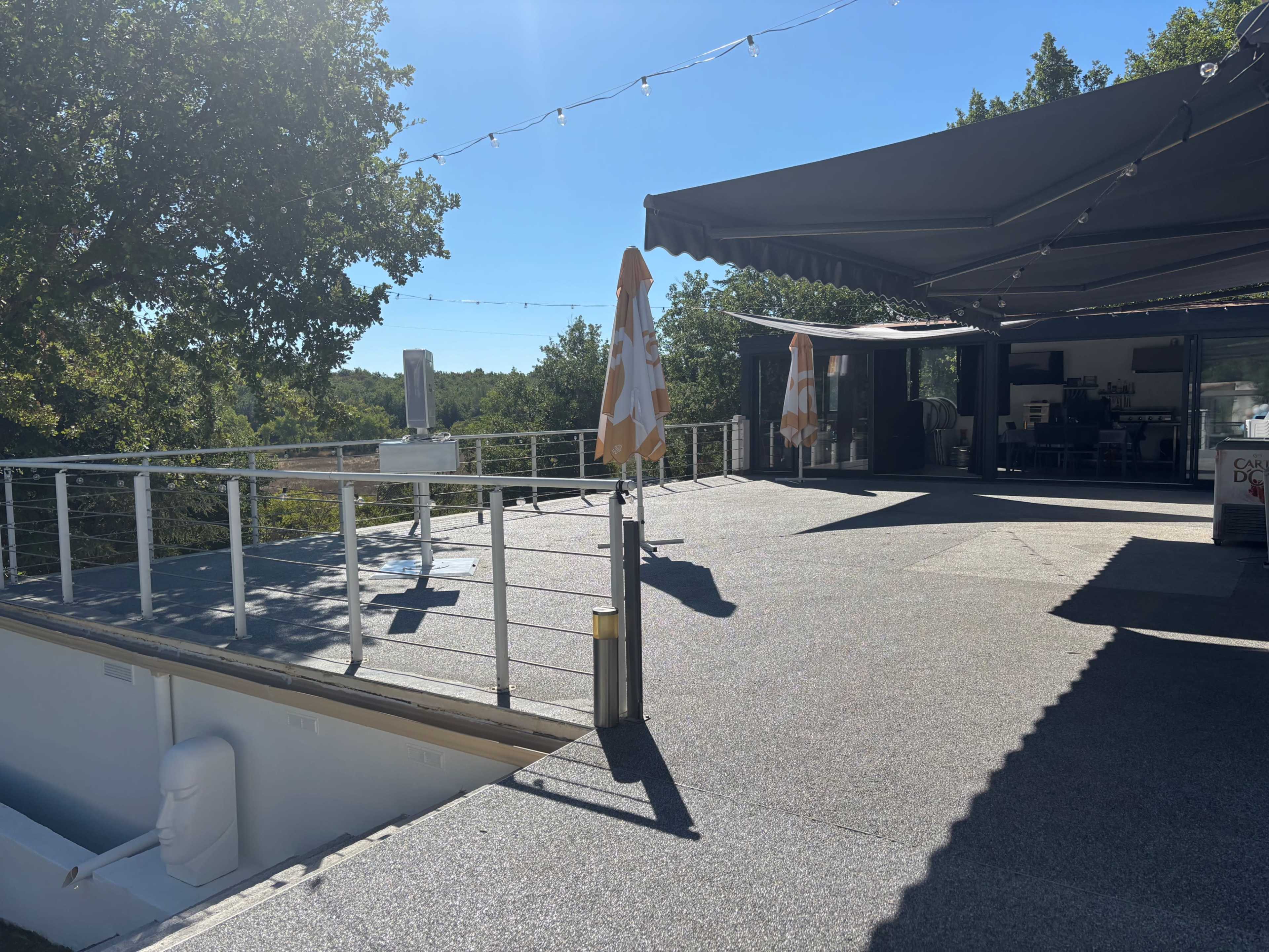 A large outdoor terrace features a flat, textured surface, two orange and white umbrellas, and a shaded area under a canopy, surrounded by greenery.