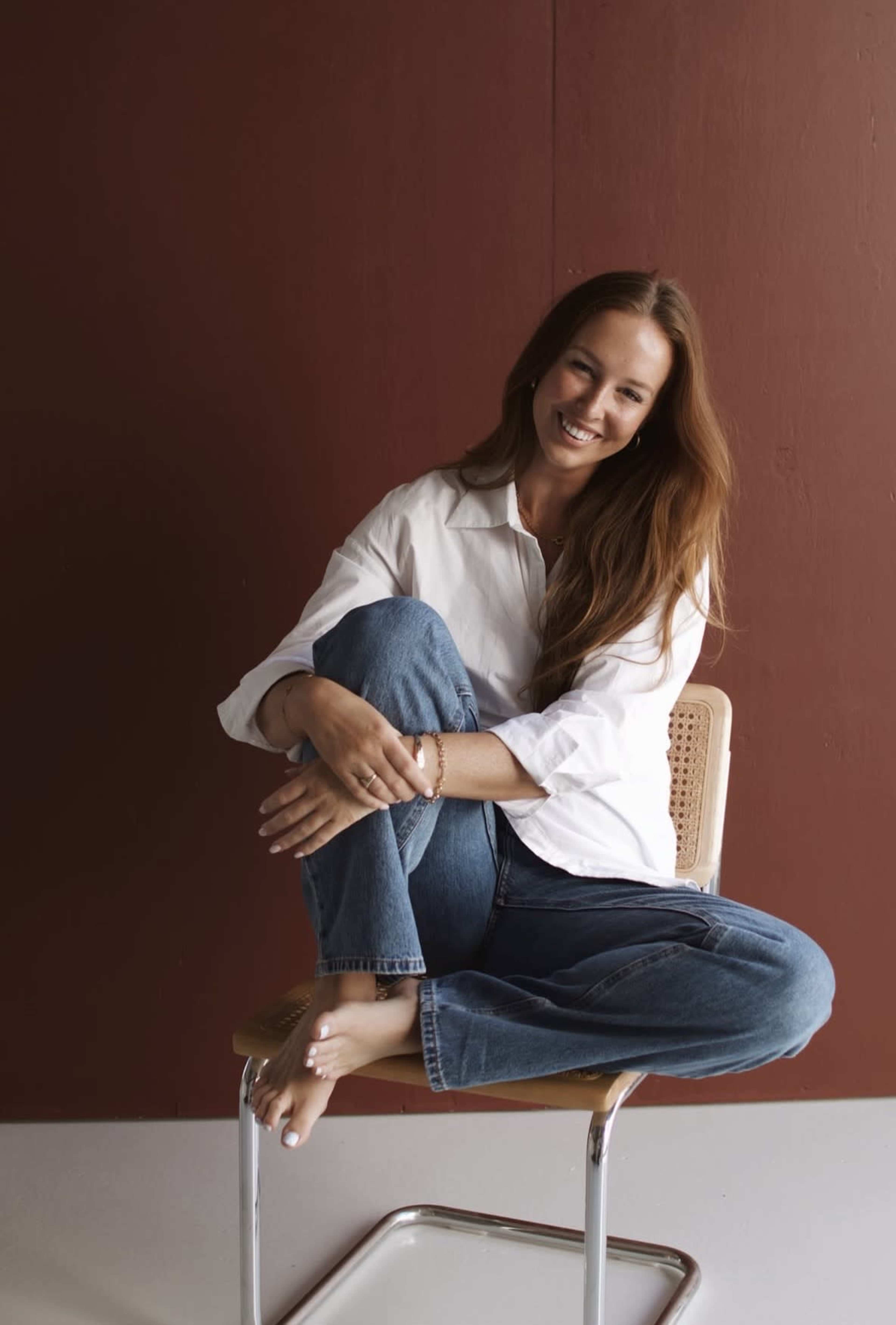 A woman with long brown hair sits cross-legged on a chair, smiling against a solid maroon wall.