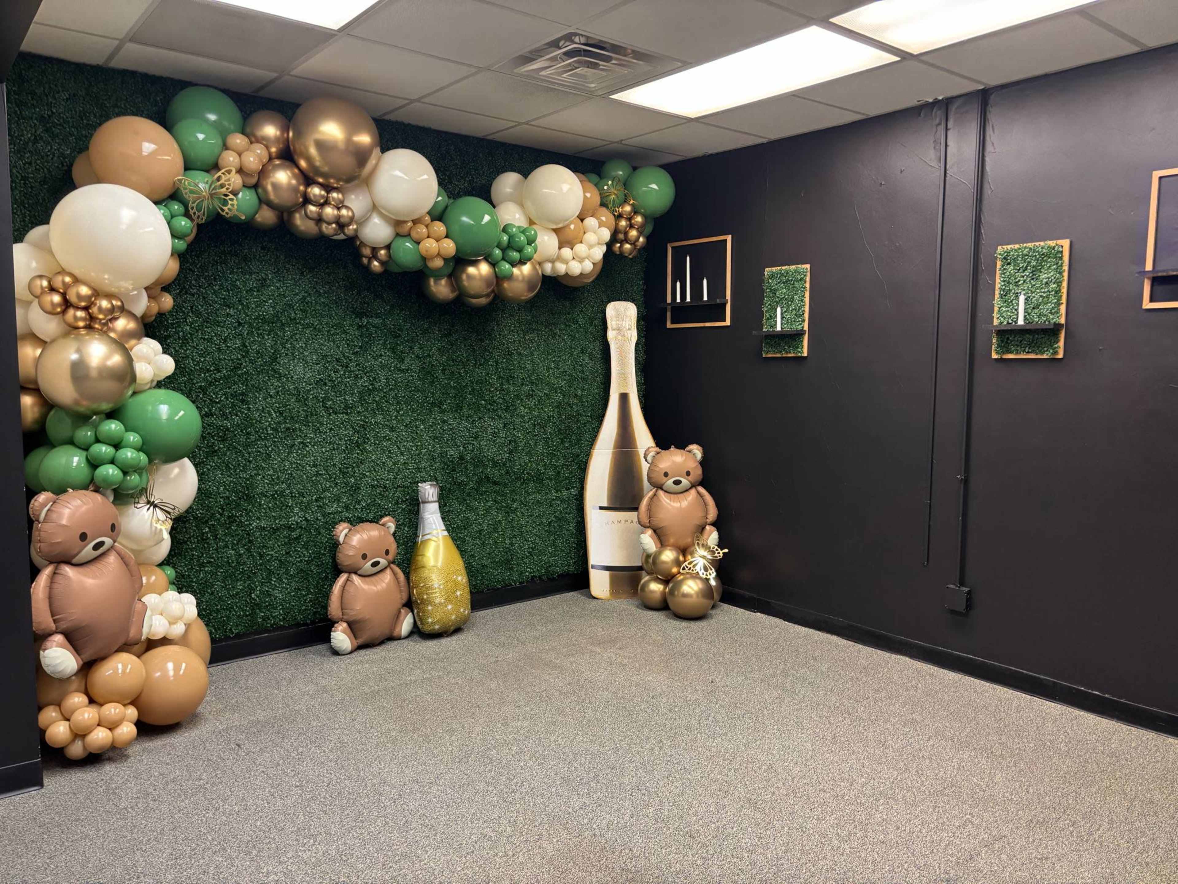 The image shows a decorated corner of a room with a green artificial grass backdrop, featuring balloon garlands in gold, white, and green, along with two teddy bear figures and a large champagne bottle display.