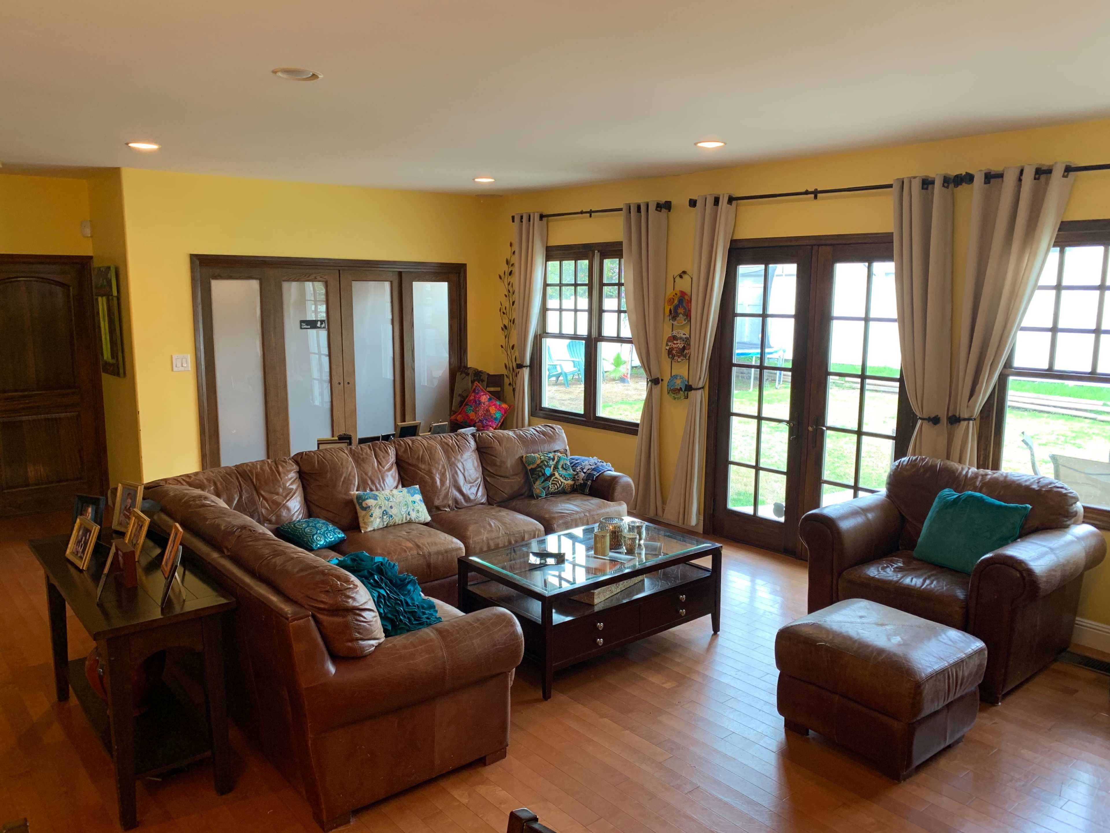 A spacious living room features a large brown leather sectional sofa around a glass coffee table, with large windows providing a view of the outdoor area.