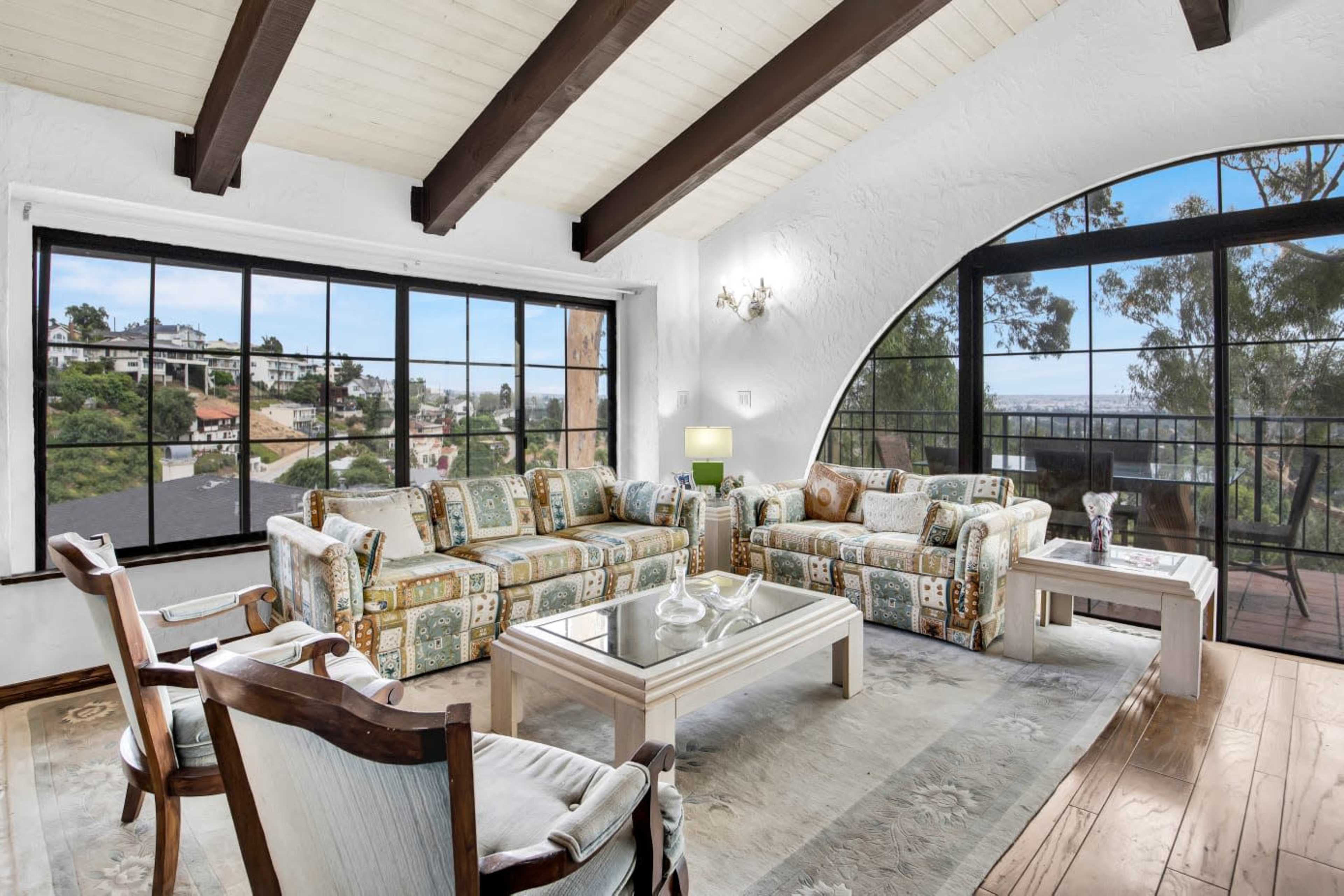 A spacious living room features arched windows with a view, wooden beams on the ceiling, and a mix of patterned furniture arranged around a glass coffee table.