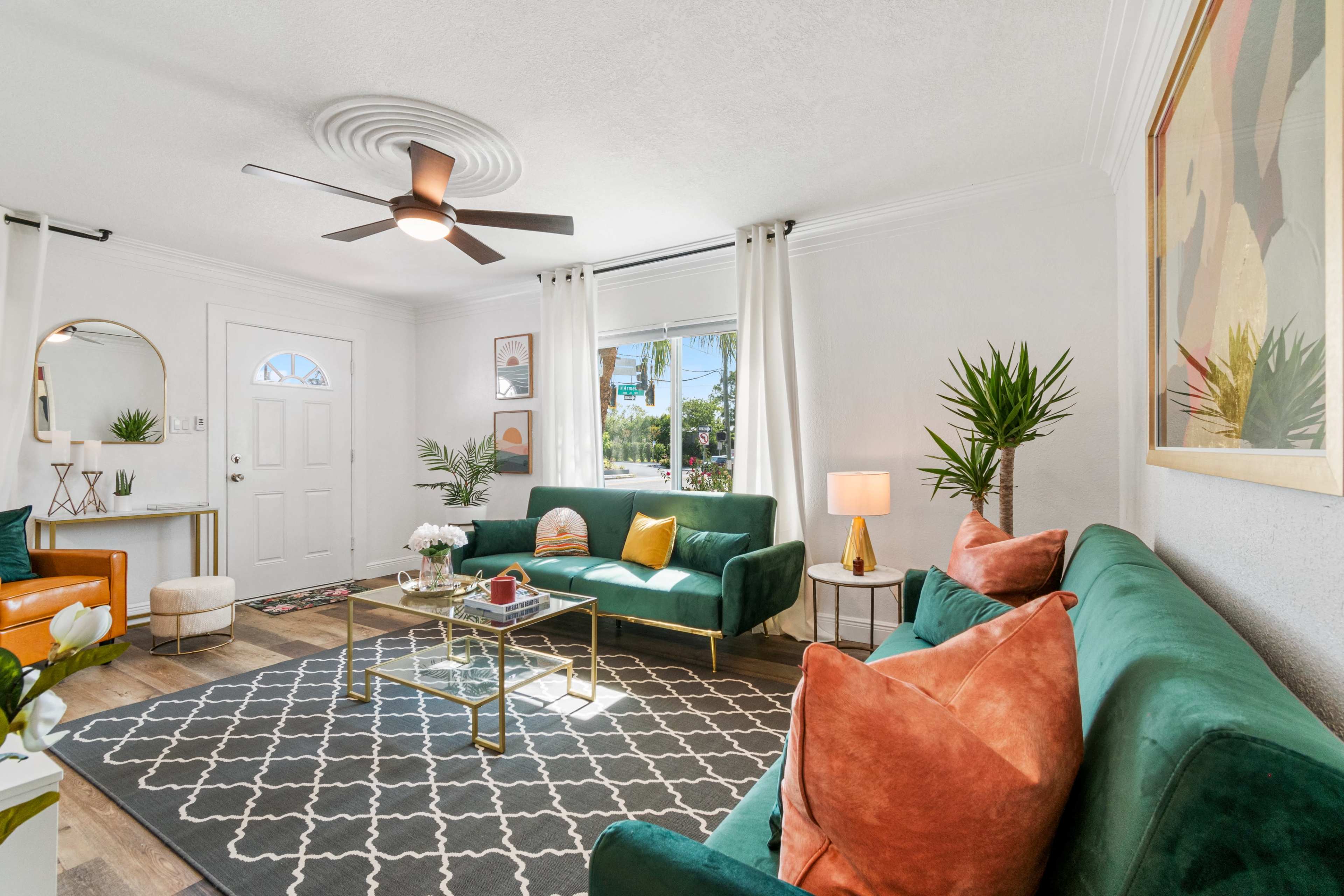 A well-decorated living room featuring green sofas, a glass coffee table, a rug, and plants by the window.