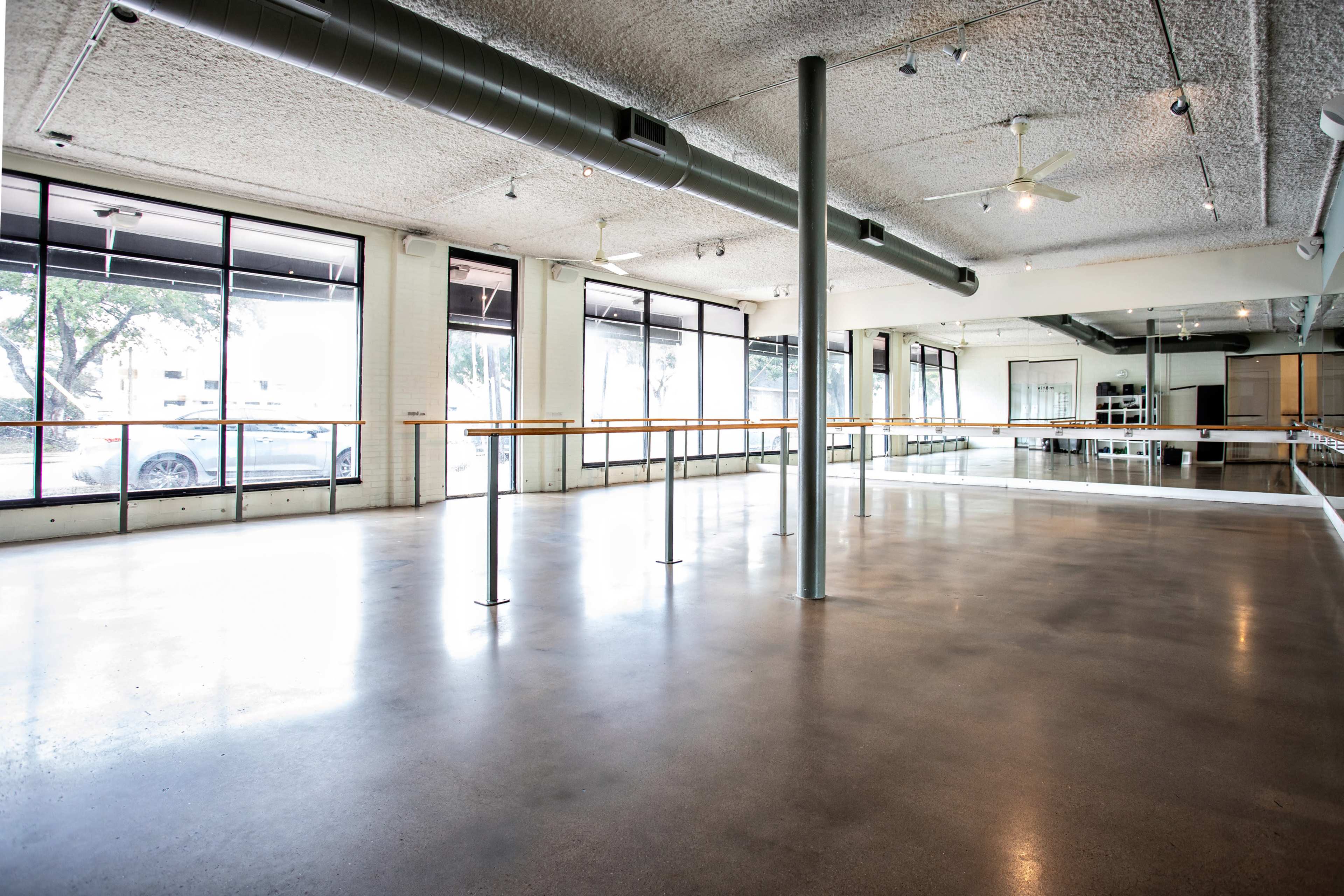An open dance studio features large windows, a mirrored wall, and a polished concrete floor.
