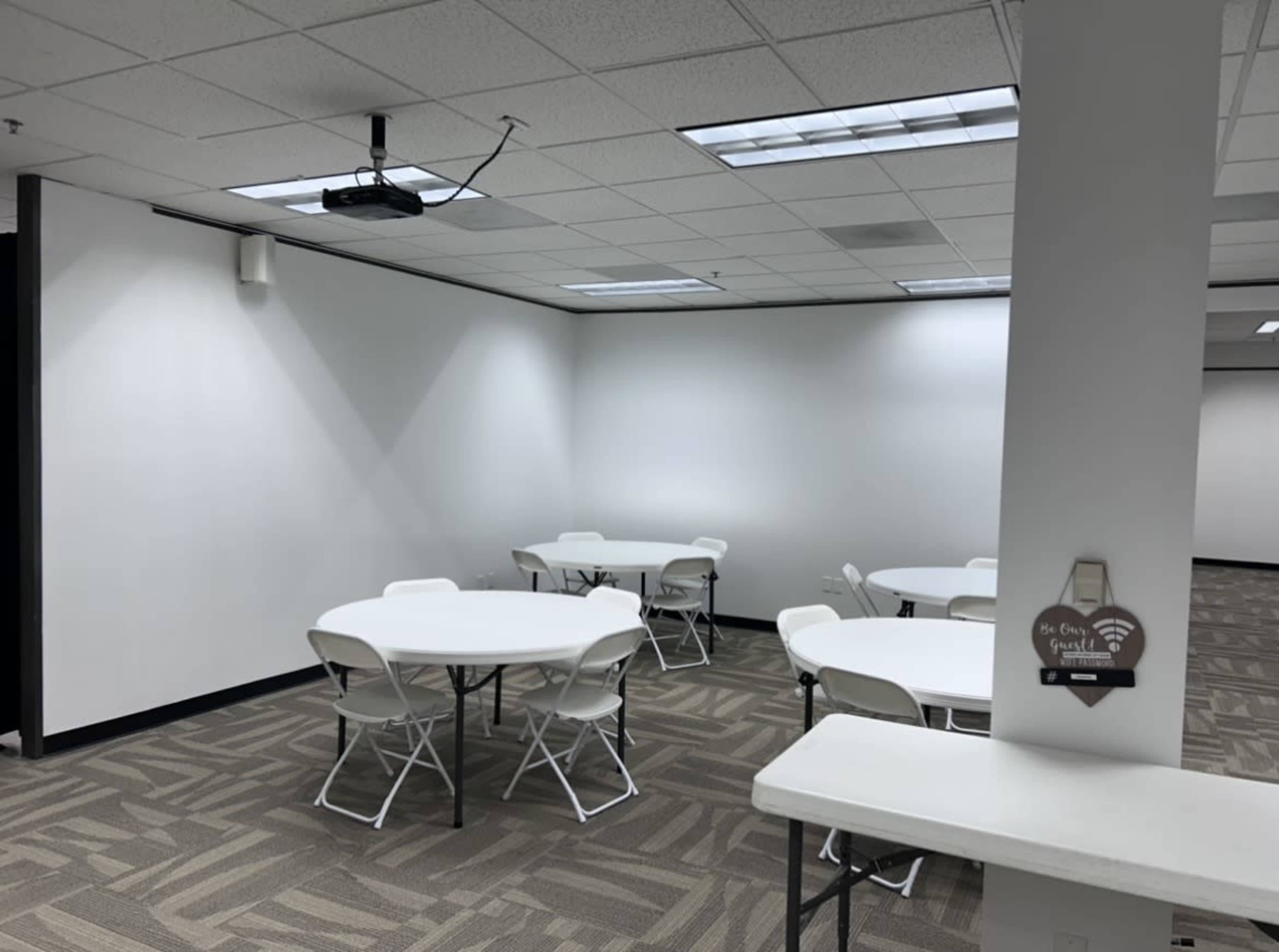 The image shows a sparsely furnished meeting room with several round tables and chairs arranged on a carpeted floor, featuring white walls and a ceiling-mounted projector.