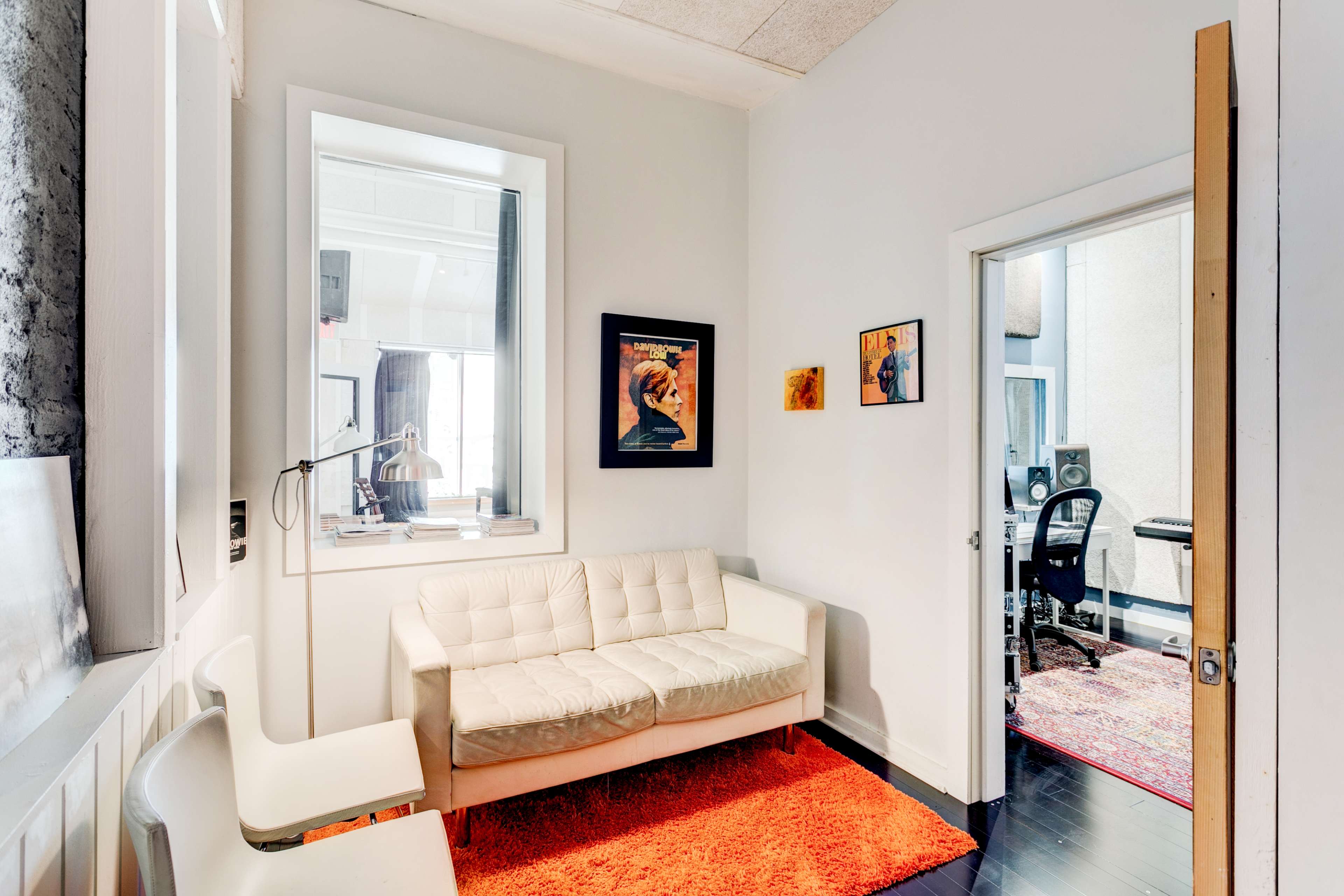 A small room with a white sofa, an orange rug, and framed artwork on the walls, leading to another room with studio equipment visible through an open door.