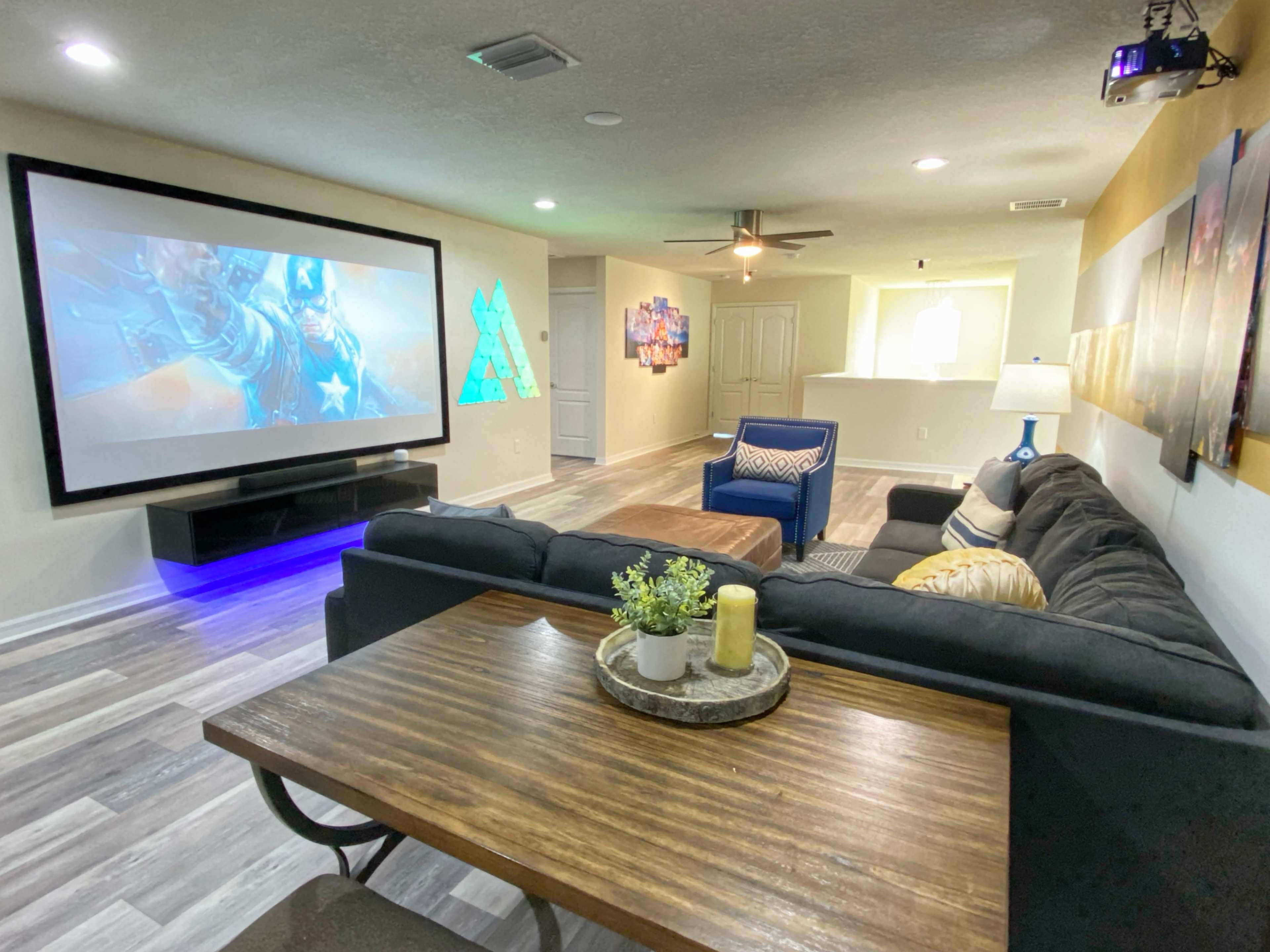 The image shows a modern living room featuring a large screen for projecting images, a sectional sofa, and a wooden dining table with decorative items.