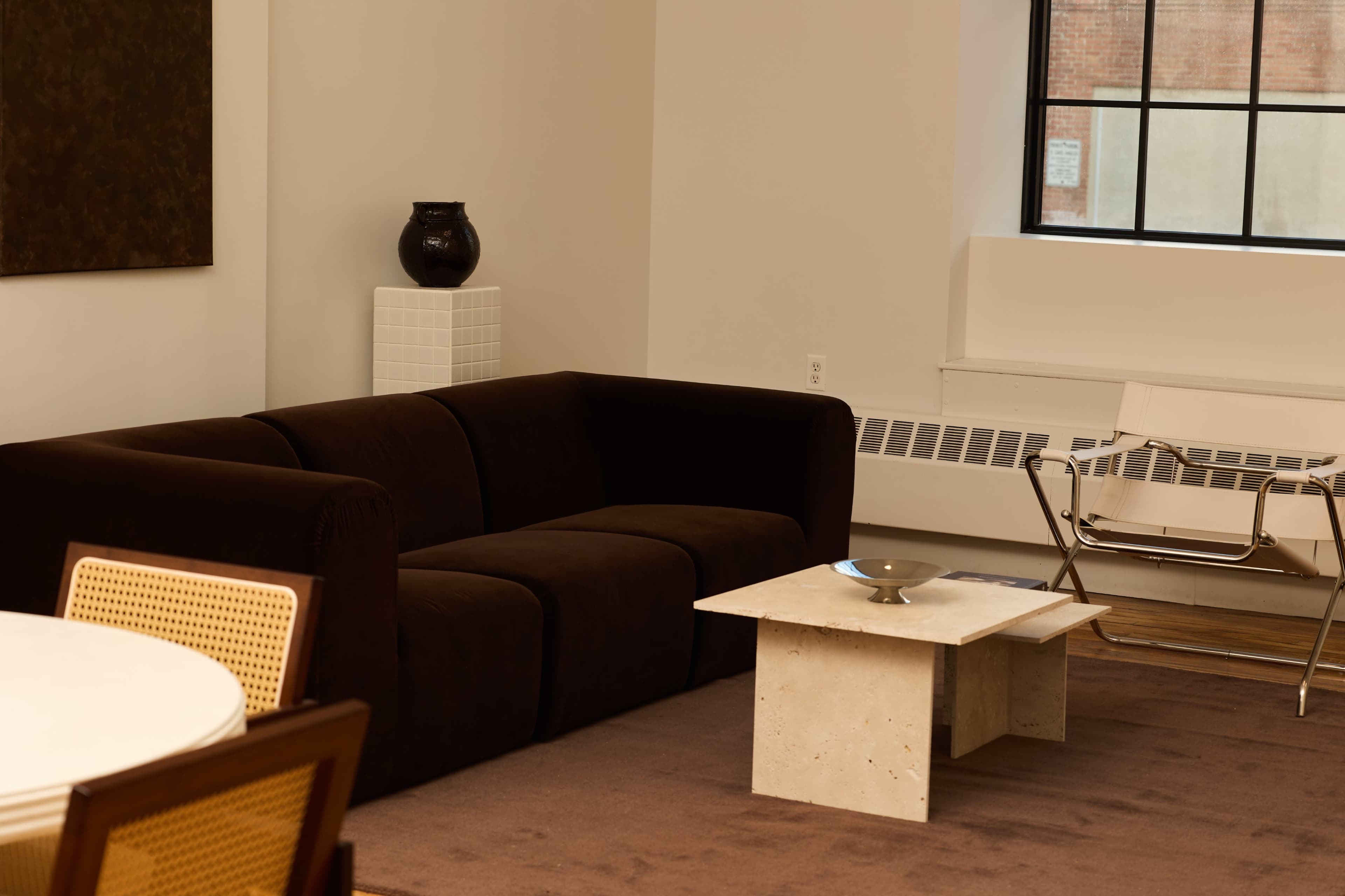 The image shows a modern living room featuring a dark sofa, a small concrete coffee table, and a chair beside a large window.