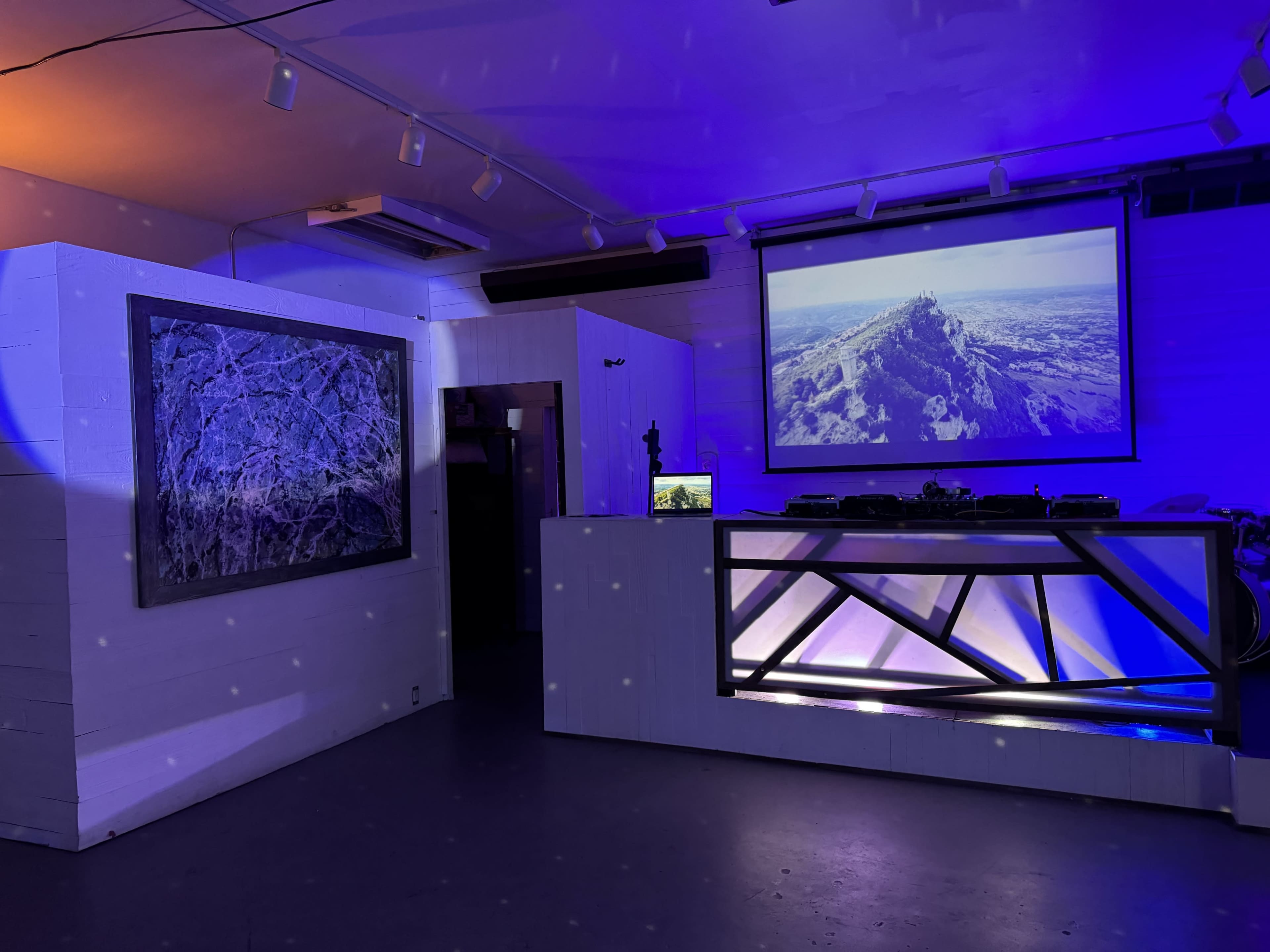 The image shows a dimly lit room featuring a DJ booth with a geometric design, an abstract artwork on the wall, and a large projection screen displaying a mountainous landscape.