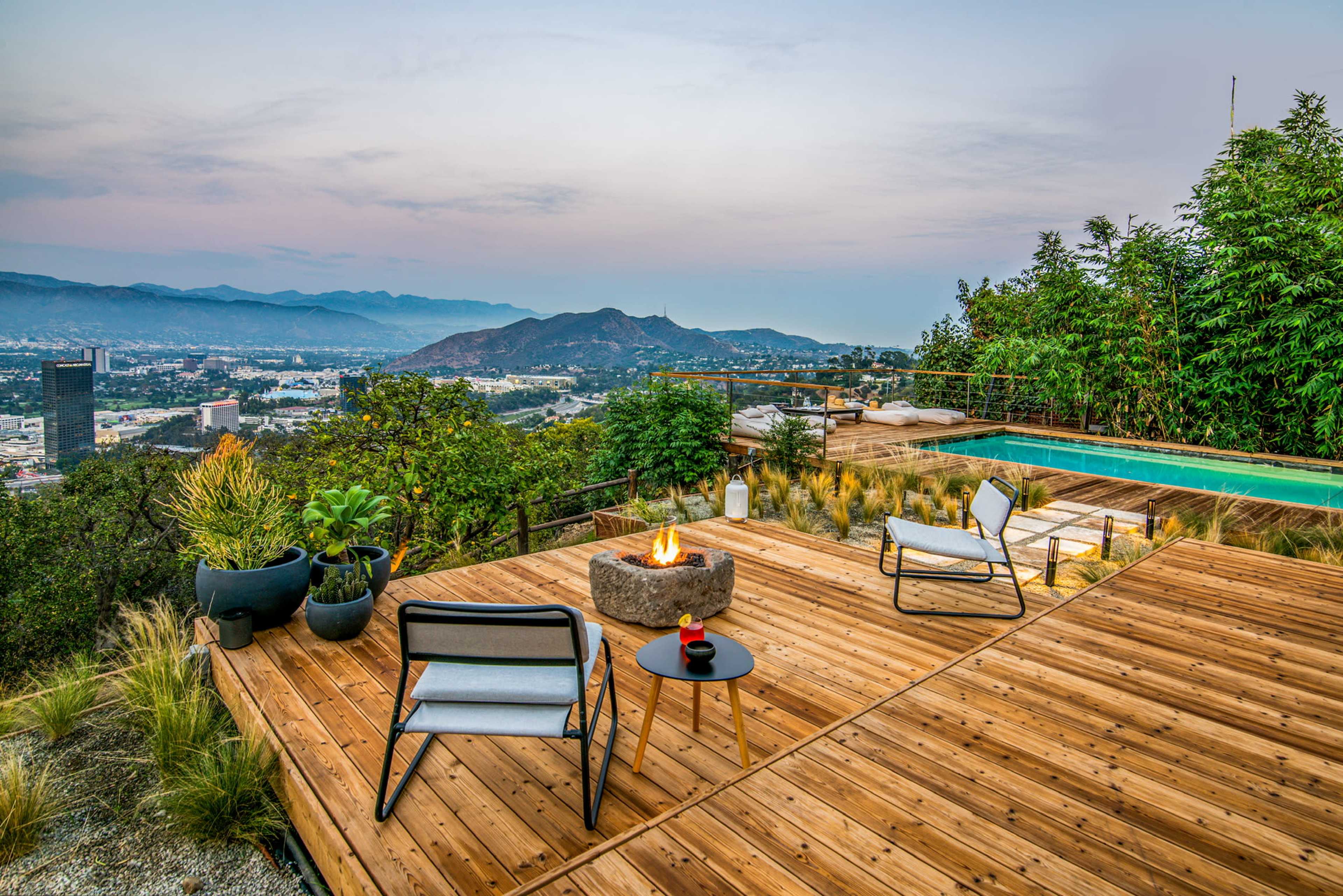 A wooden deck features two chairs and a small table near a fire pit, overlooking a valley with mountains and a swimming pool in the background.