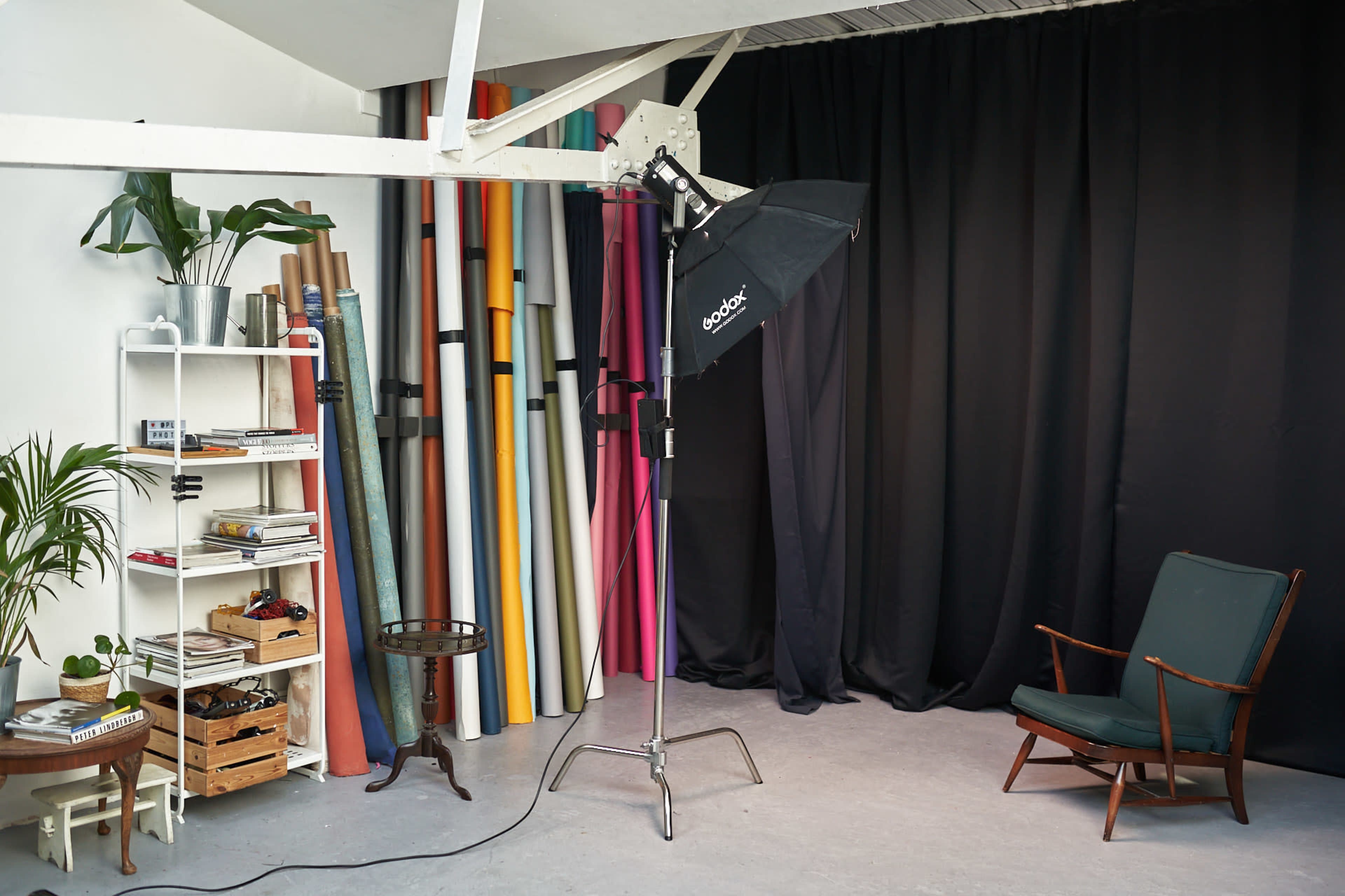 A photography studio setup with a softbox light, a green chair, a small table, and colorful backdrops arranged along one wall.