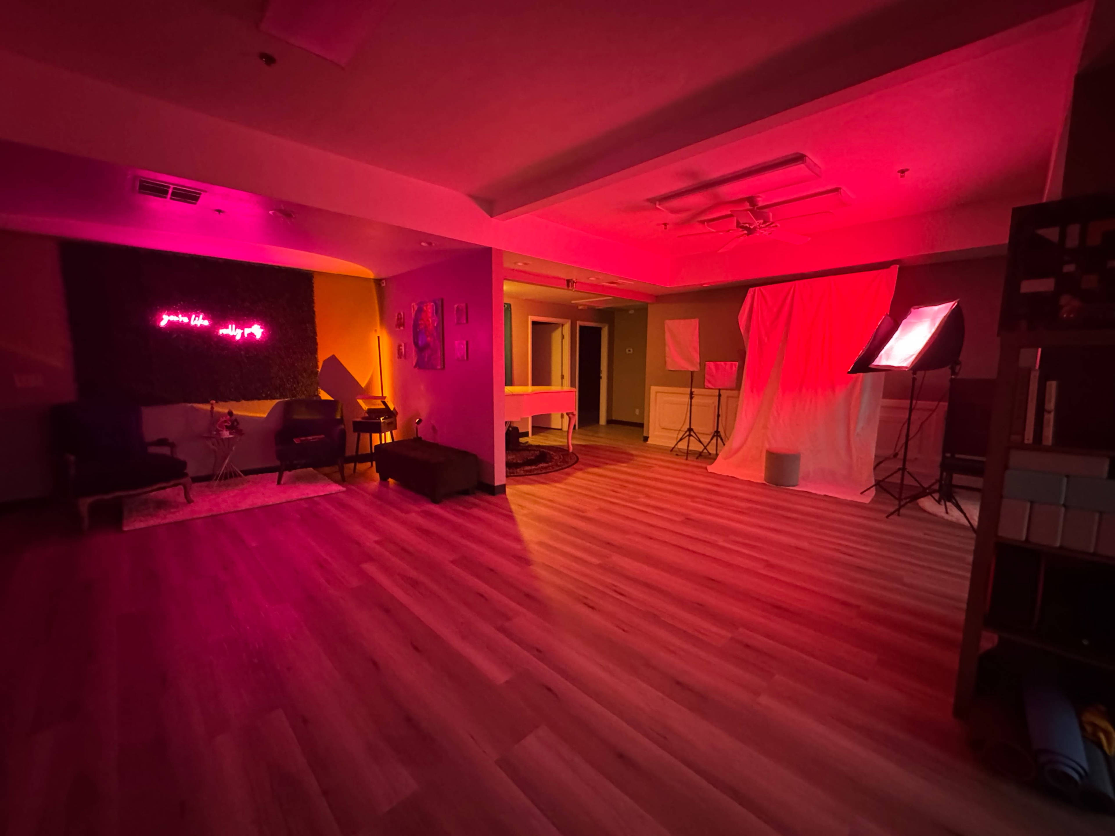 The interior of a dimly lit room is illuminated with pink lighting, featuring various furniture pieces and a backdrop draped in white fabric.