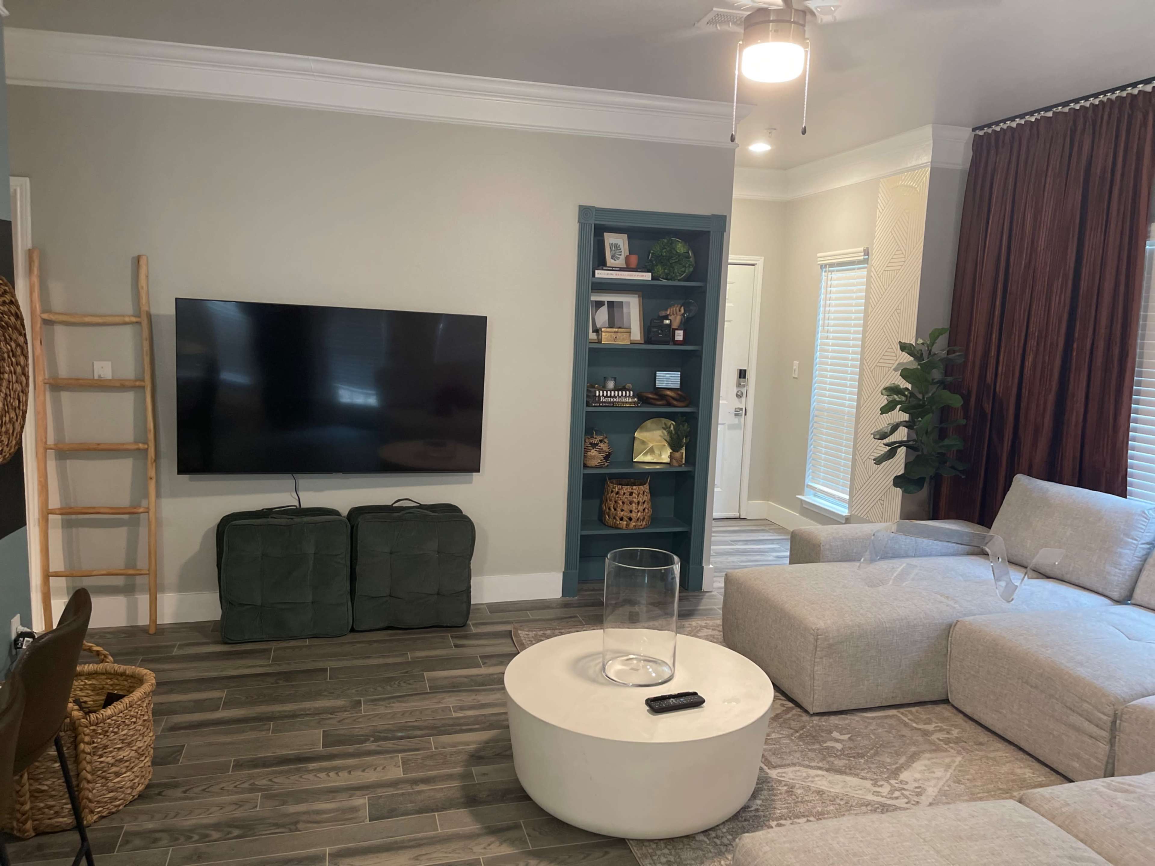 A modern living room features a grey sectional sofa, a round coffee table, a large television mounted on the wall, and a green bookshelf filled with decor.
