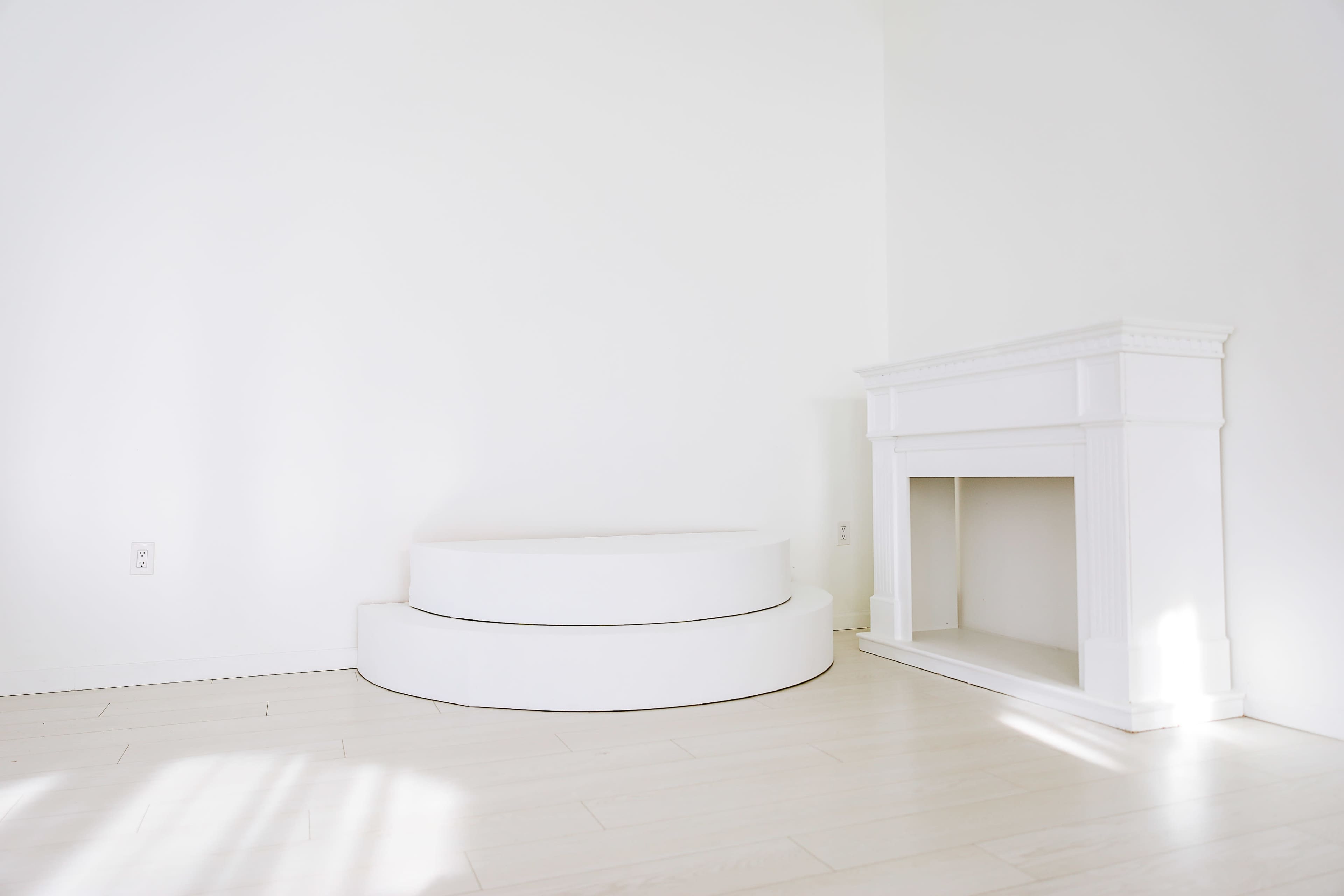 The image depicts a minimalistic room with a white curved seating platform and a white fireplace against a plain wall.