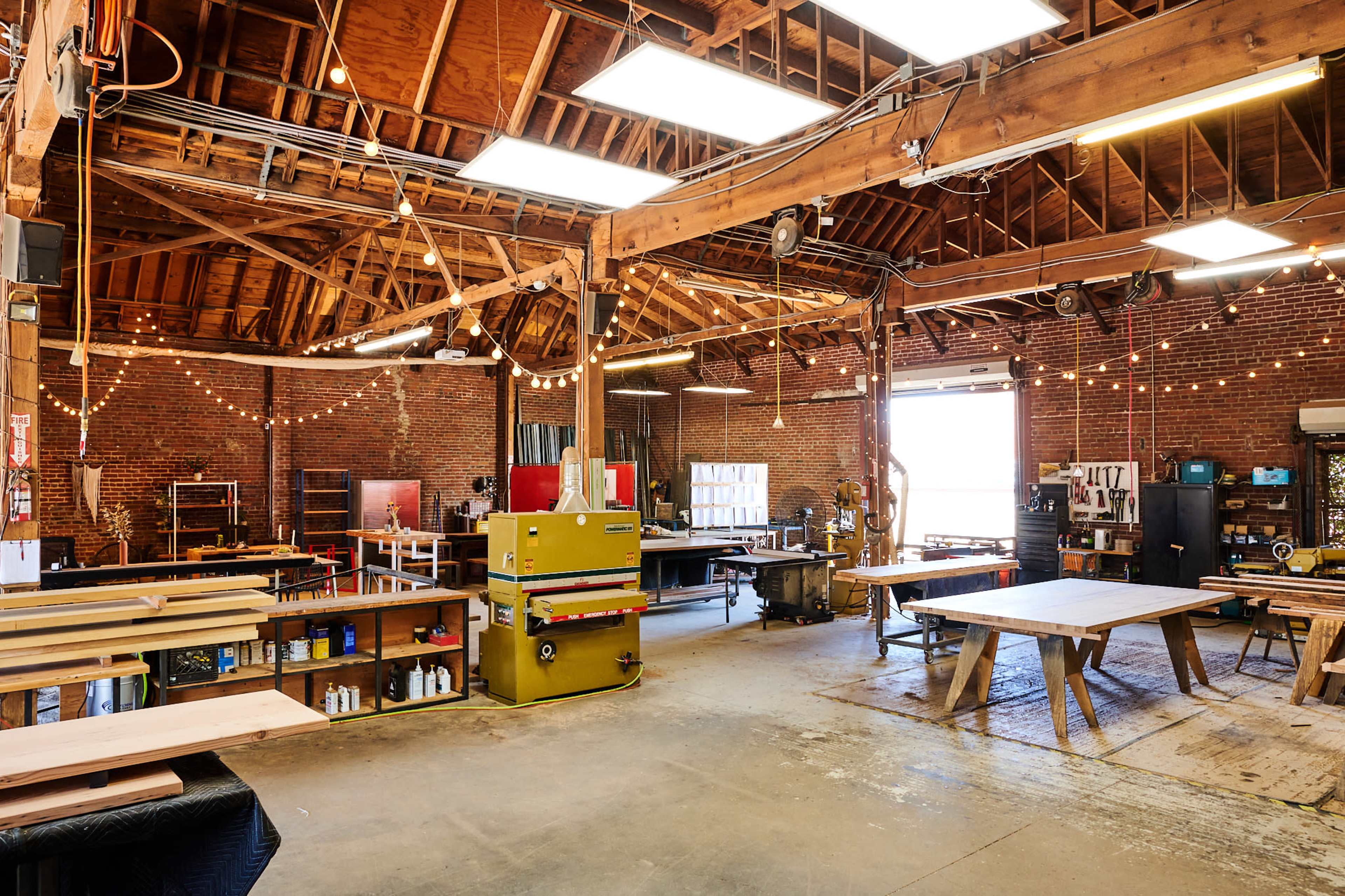 A well-lit woodworking shop featuring exposed brick walls, multiple worktables, and shelves stocked with tools and materials.
