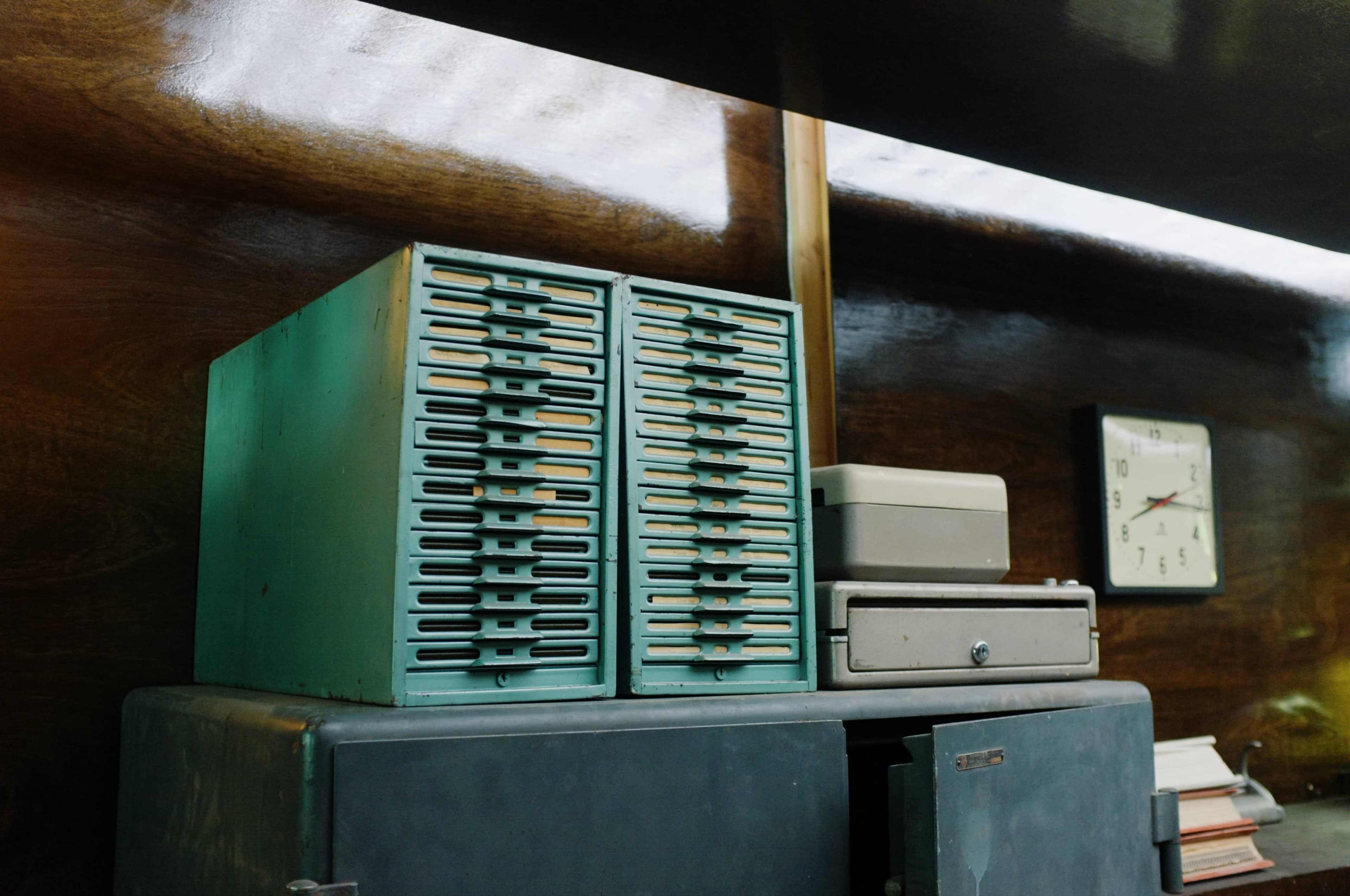 A wooden shelf with two blue vertical filing cabinets stacked on top, alongside a small light gray box and a clock mounted on the wall.