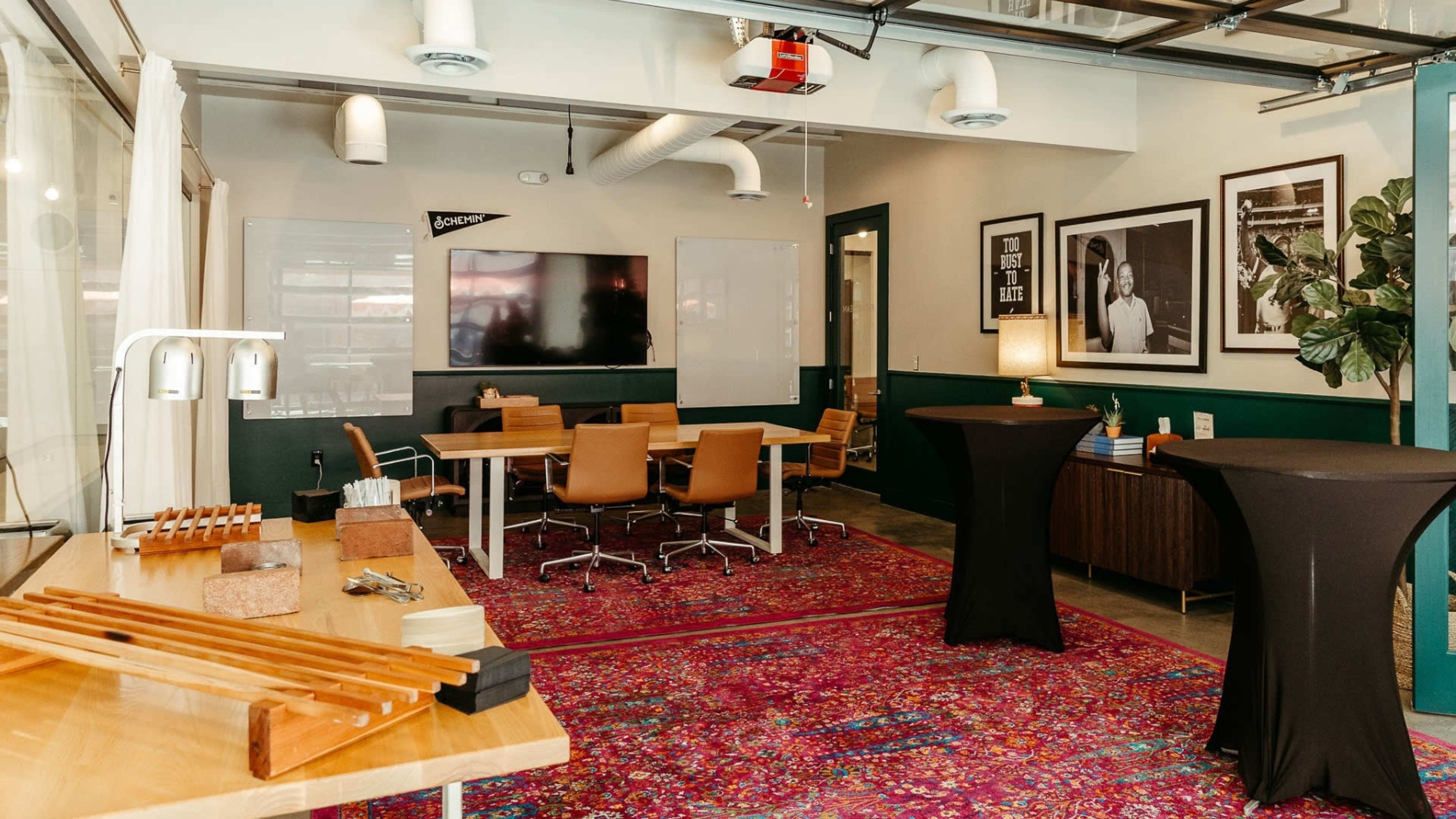 The image shows a modern workspace with a conference area featuring a large screen, wooden tables, and colorful rugs, set up with chairs and decorative wall art.