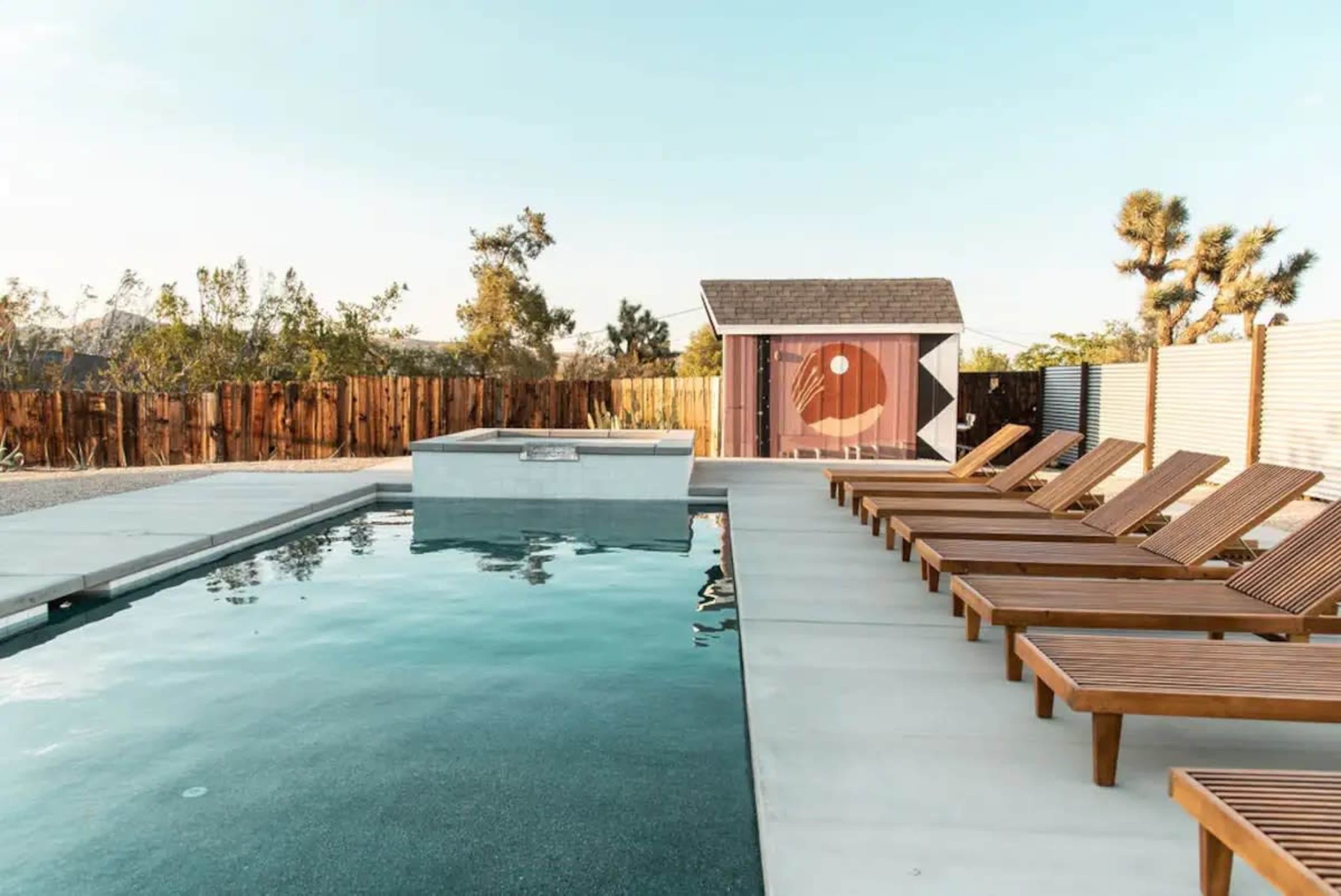 A modern outdoor pool area with wooden lounge chairs and a small shed surrounded by a wooden fence and trees.