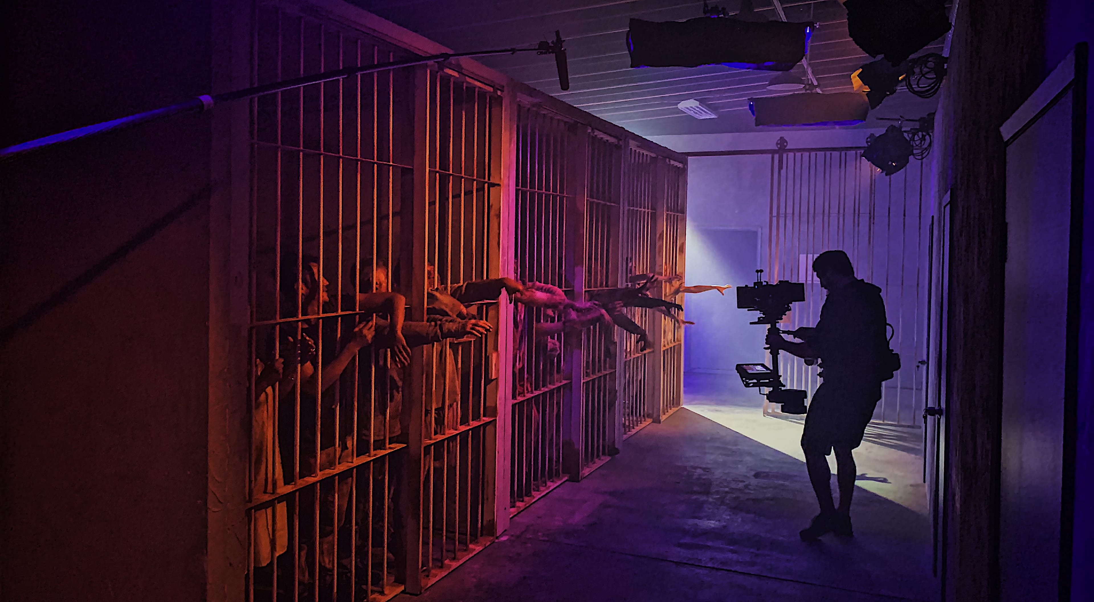 A film crew captures a scene in a dimly lit corridor of a prison set, with actors reaching through bars in the background while a camera operator films.