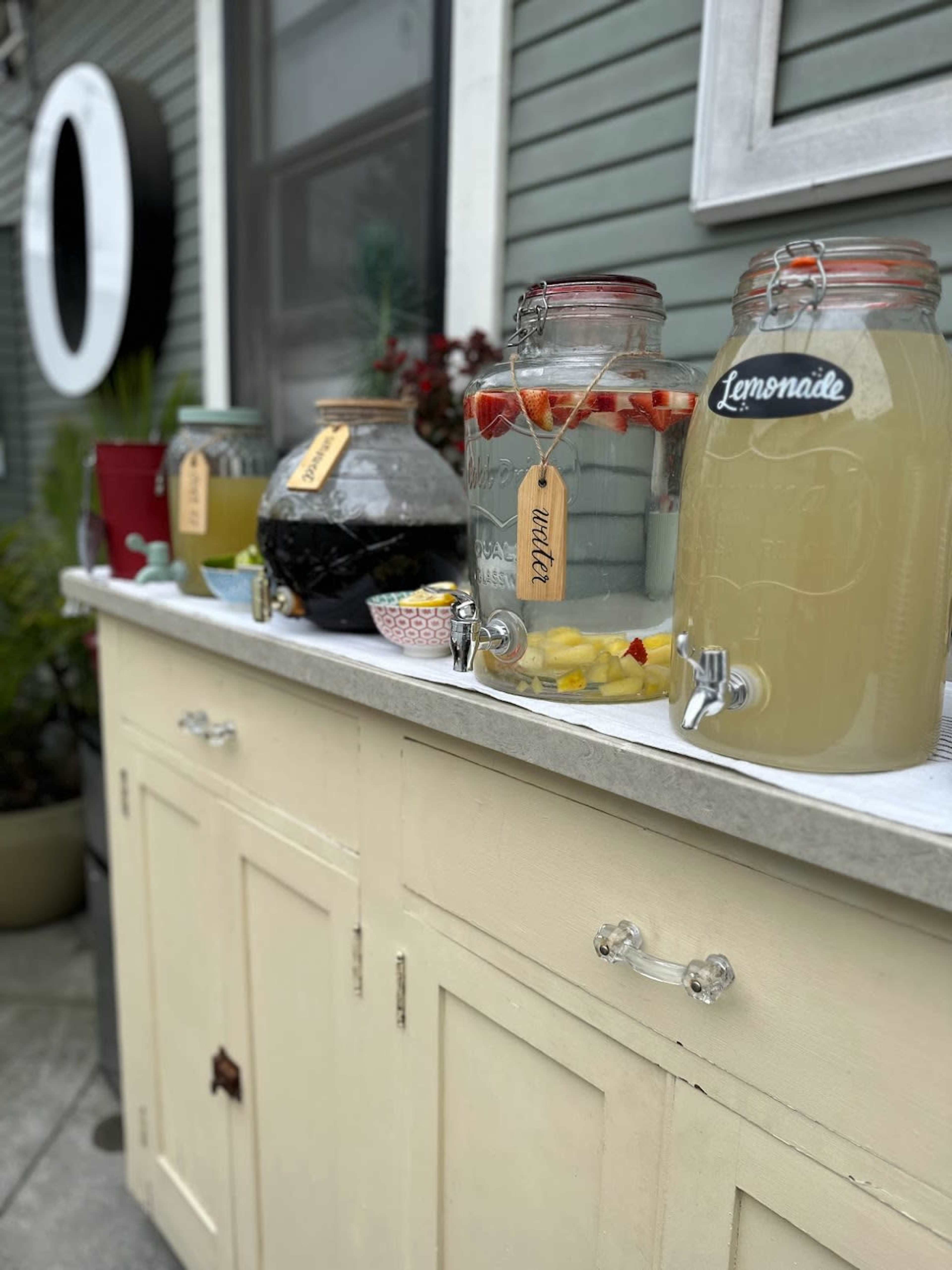 A beverage station with several glass dispensers containing lemonade, infused water, and other drinks, arranged on a cabinet.