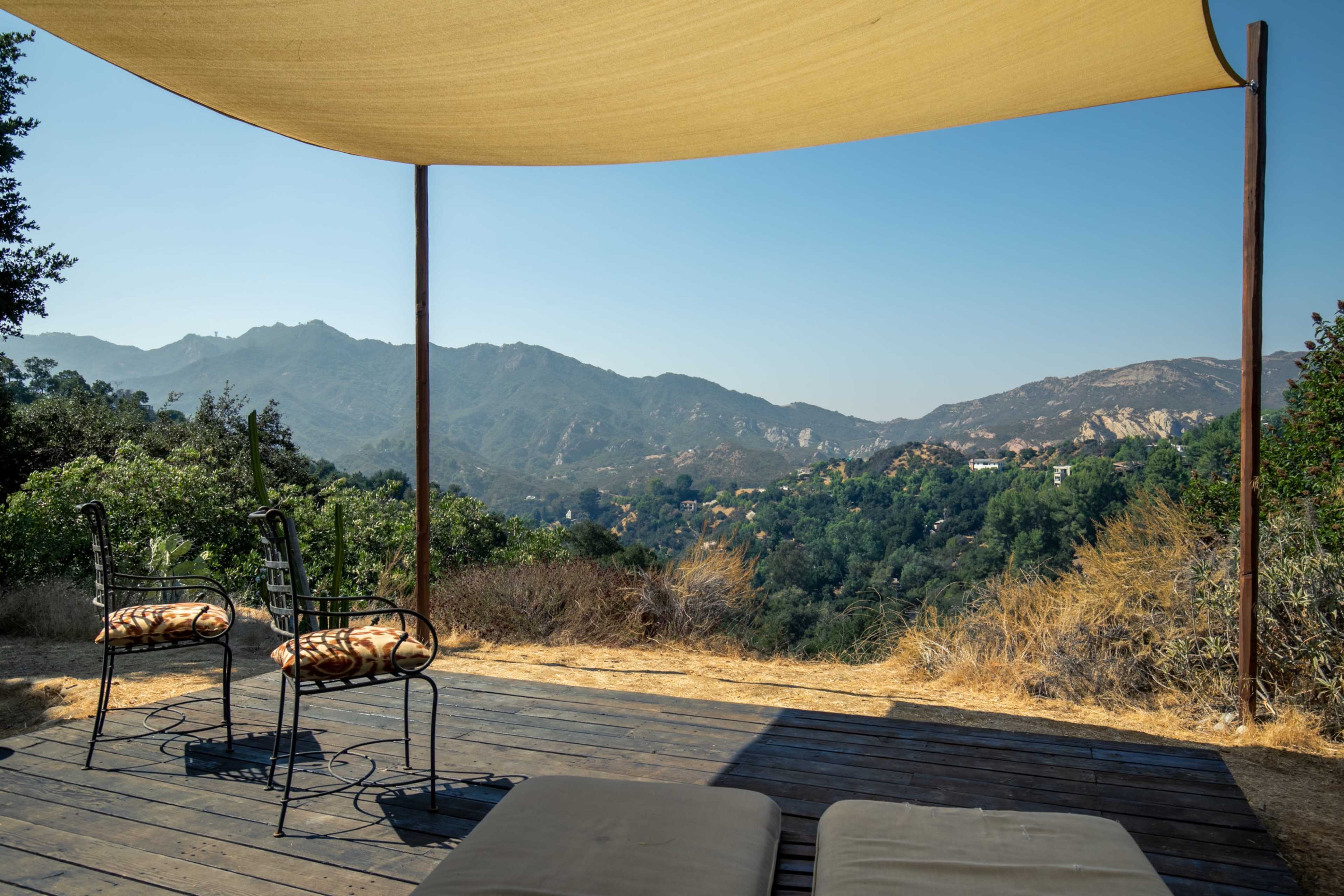 A shaded outdoor seating area with two chairs overlooks rolling hills and mountains under a clear blue sky.