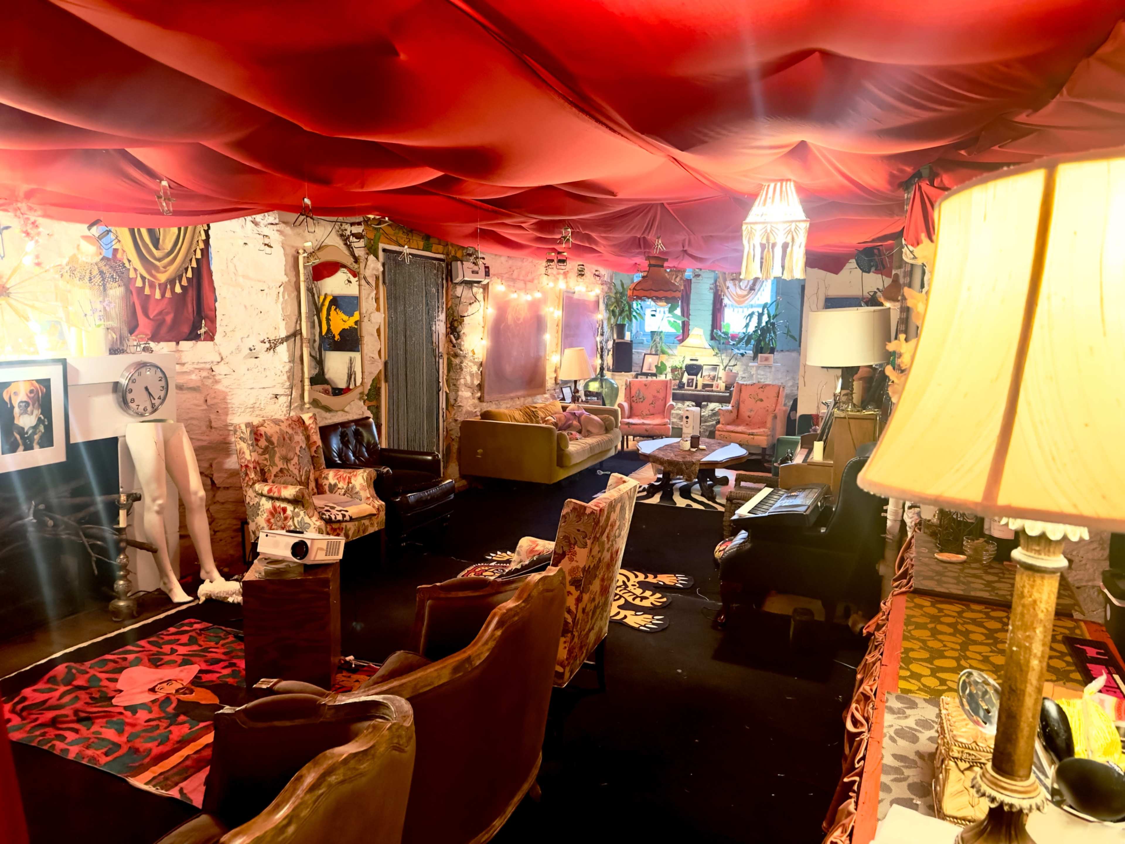 The image depicts a cozy living space featuring eclectic seating, a red draped ceiling, and various decorative items against a textured wall.