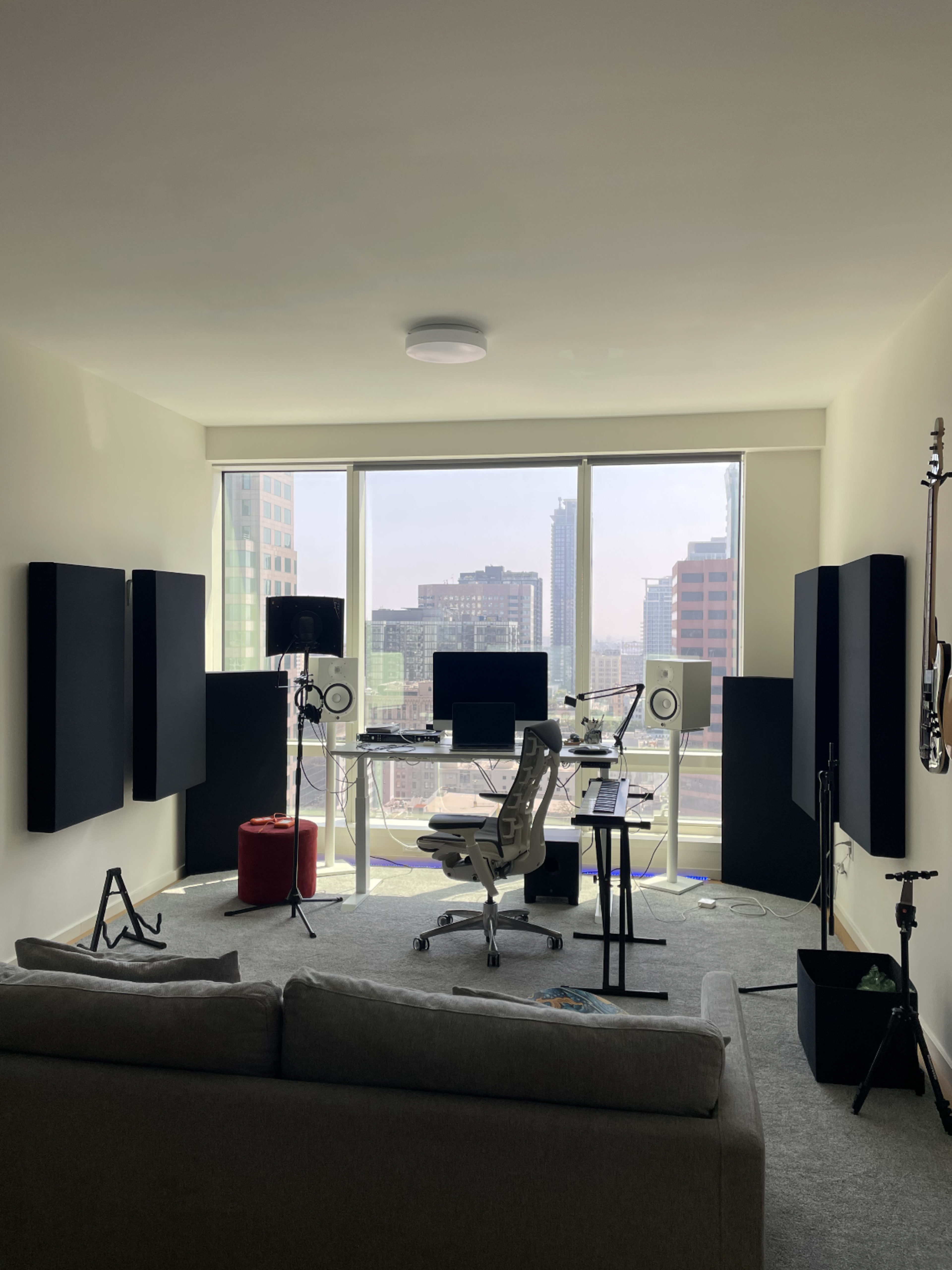 The image shows a modern recording studio setup with audio equipment, a computer desk, and large windows revealing a cityscape.