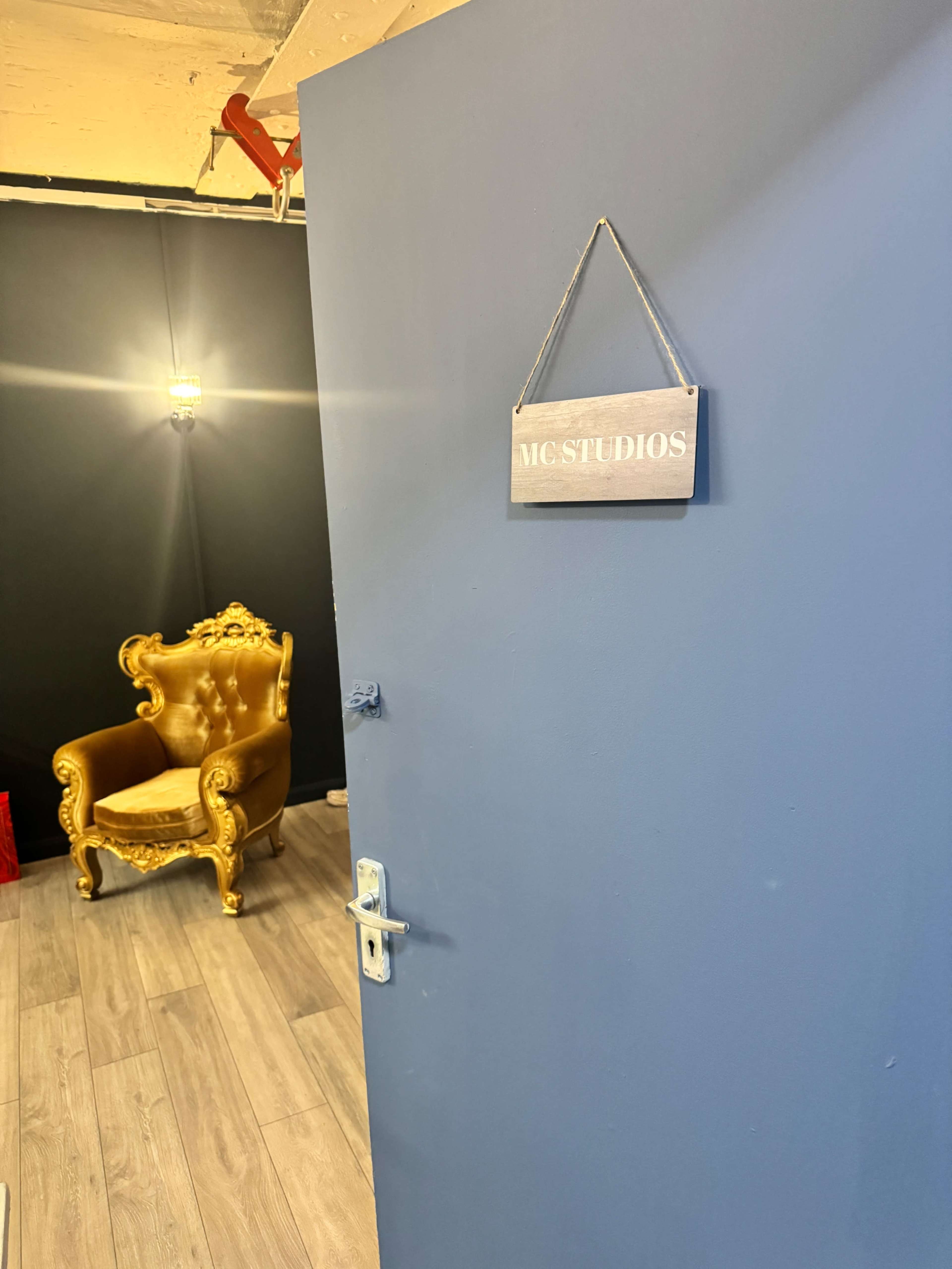 A blue door with a sign that reads "MC STUDIOS" stands ajar, revealing a decorative golden chair in a dimly lit room.