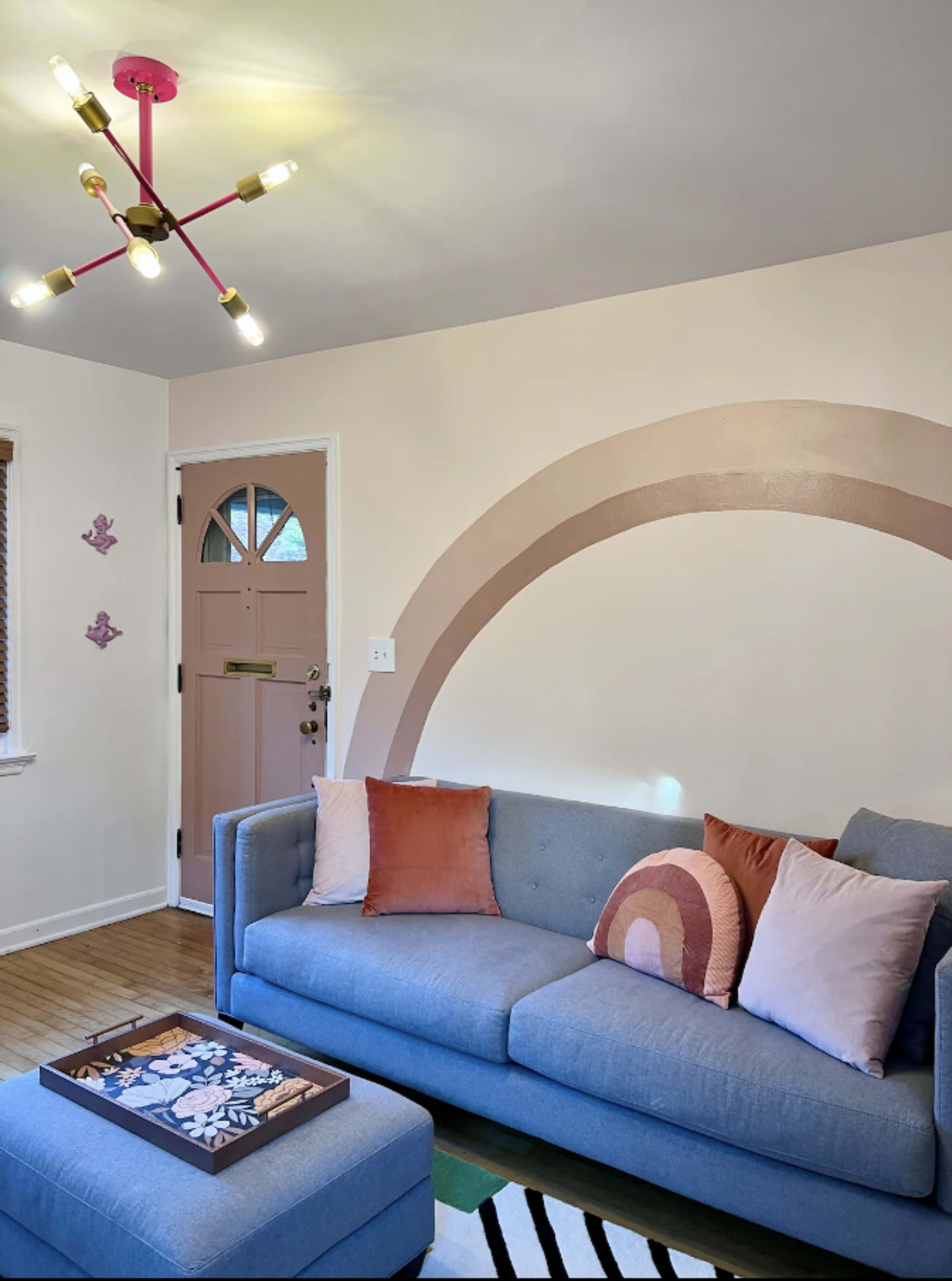 A modern living room with a blue sofa adorned with decorative pillows, a coffee table featuring a decorative tray, and a pastel-colored arch painted on the wall above.