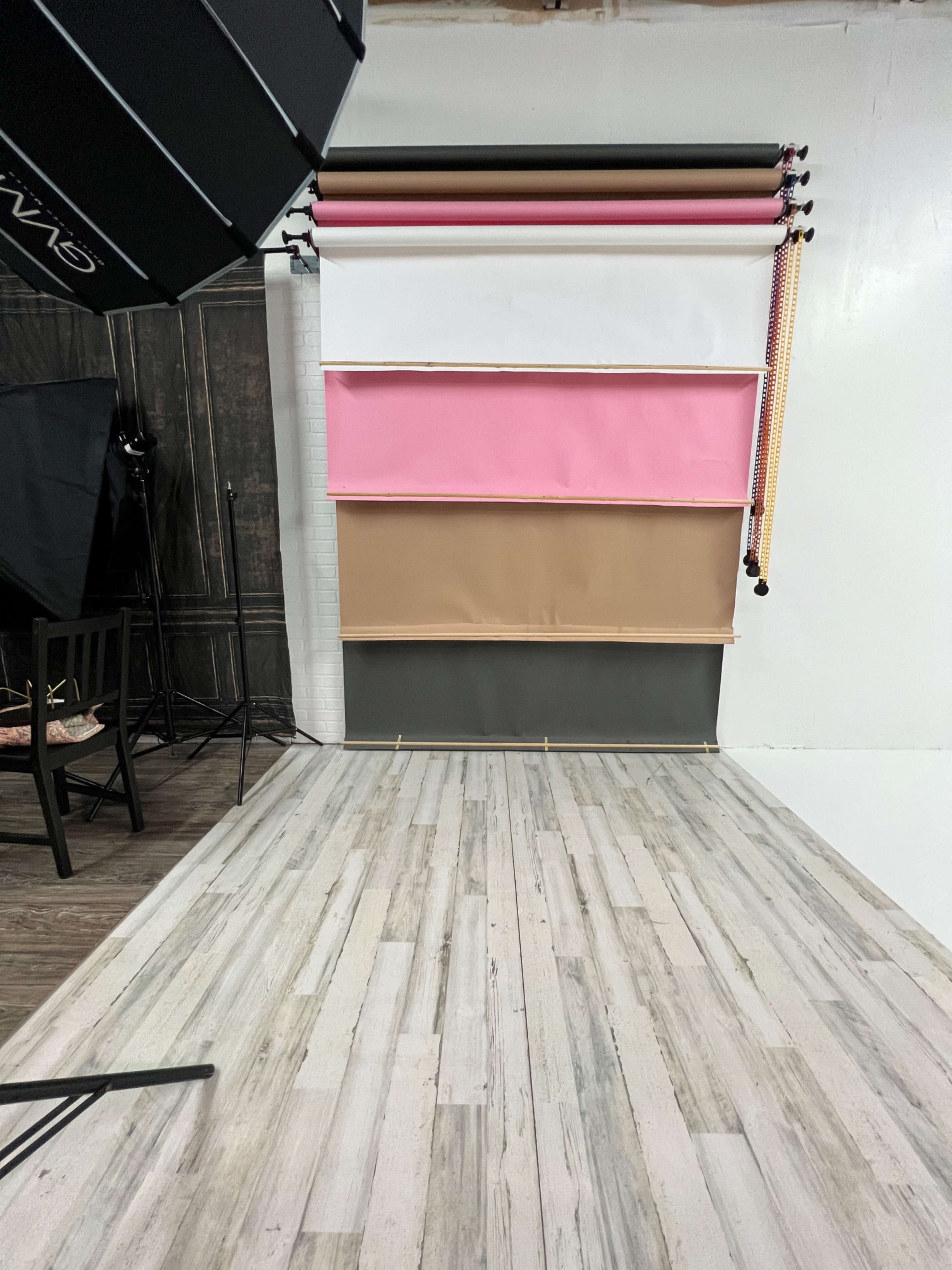 A photography studio setup with various colored backdrops rolled up against a wall and a wooden floor.