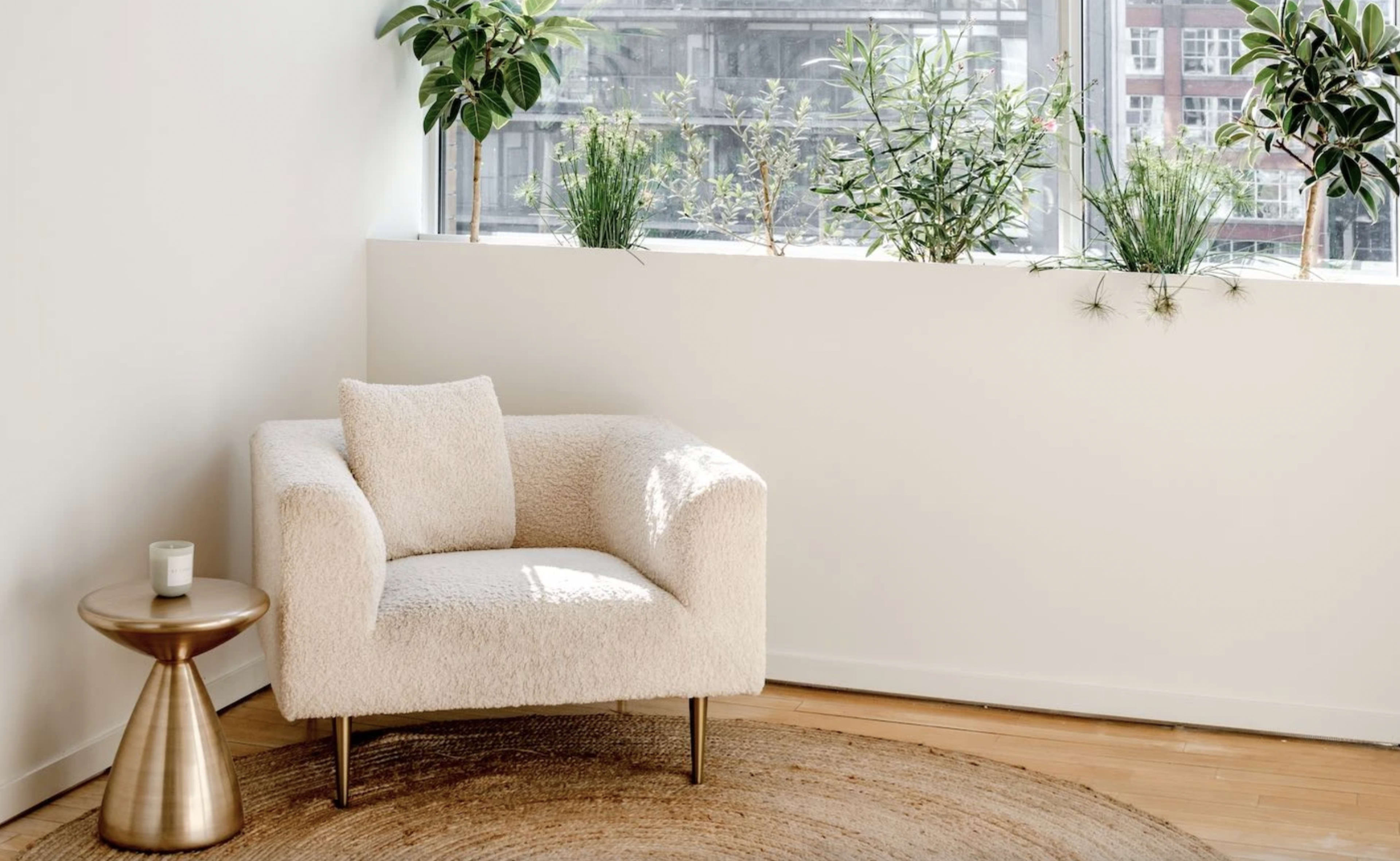 A cream-colored armchair sits beside a small round table in a bright room filled with greenery along a large window sill.