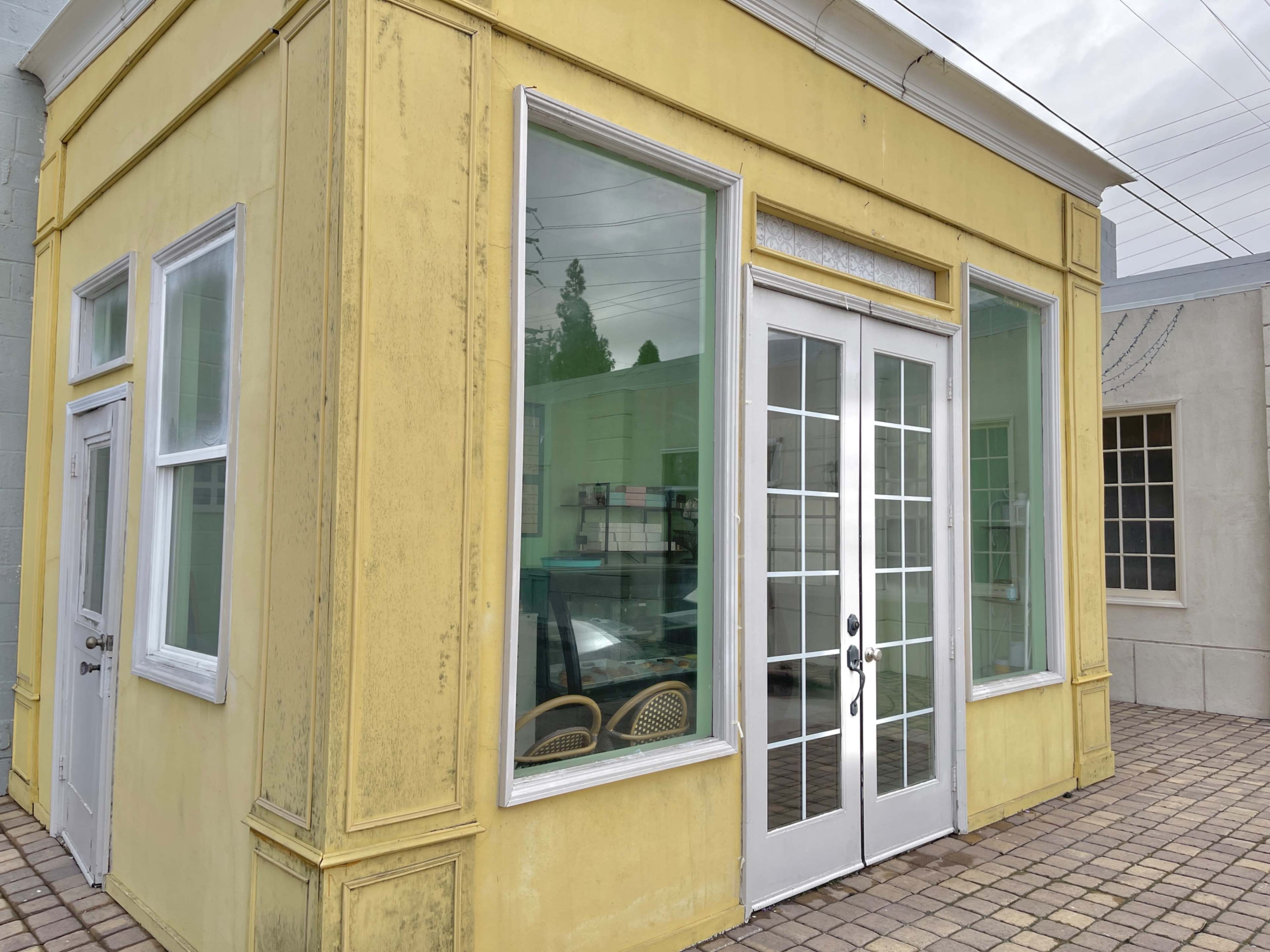 A yellow, one-story building with large glass windows and double doors, situated on a paved surface.