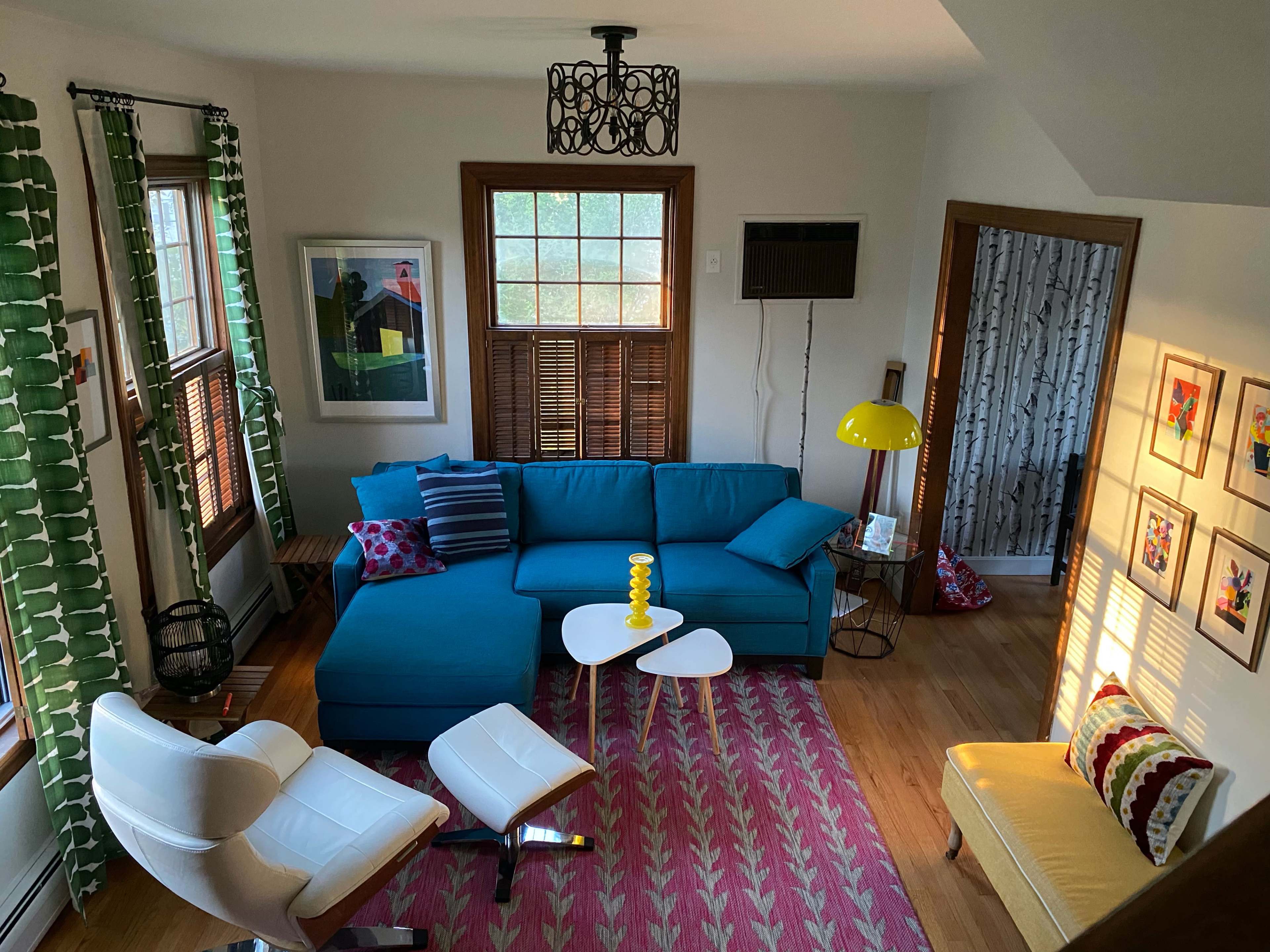 A cozy living room with a turquoise sectional sofa, a white coffee table, and a yellow floor lamp, complemented by colorful artwork on the walls and sunlight streaming through the windows.