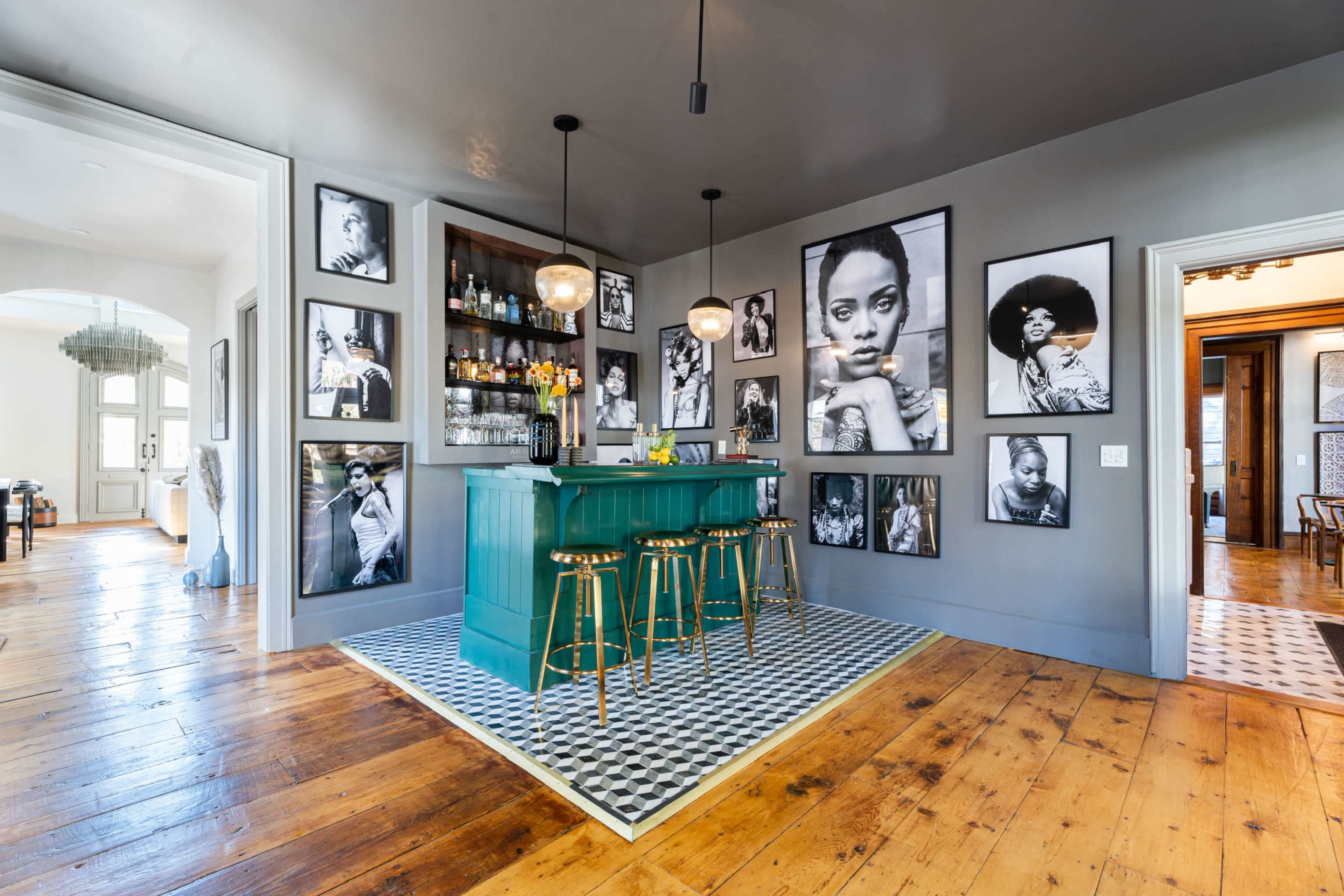 A home bar area featuring a turquoise bar with gold stools, surrounded by black-and-white portrait photographs on the walls, set against a backdrop of wooden flooring and patterned tiles.