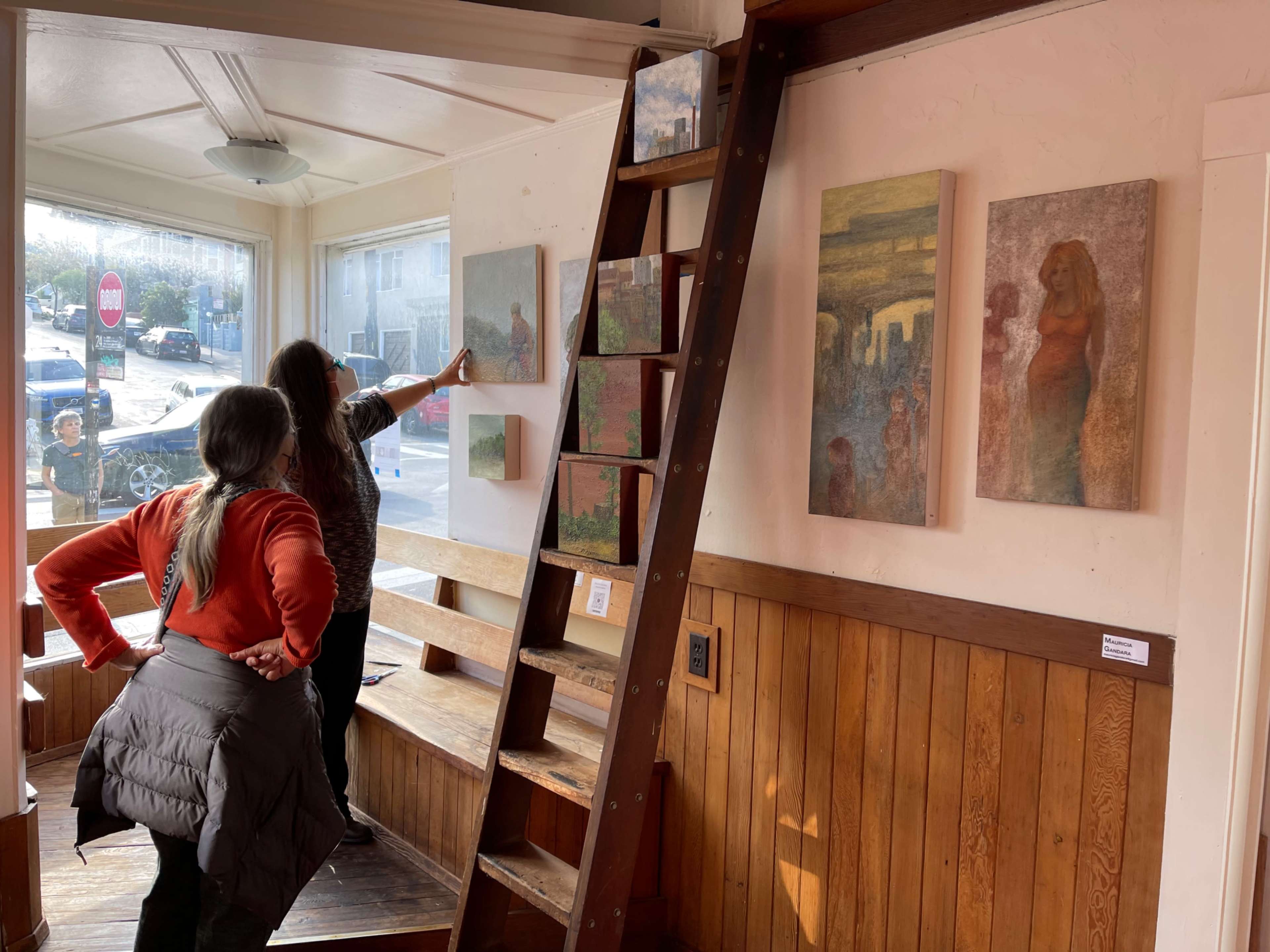 Two women examine artwork on the walls of a gallery, while a wooden ladder leans against one wall and a street view is visible through the window.