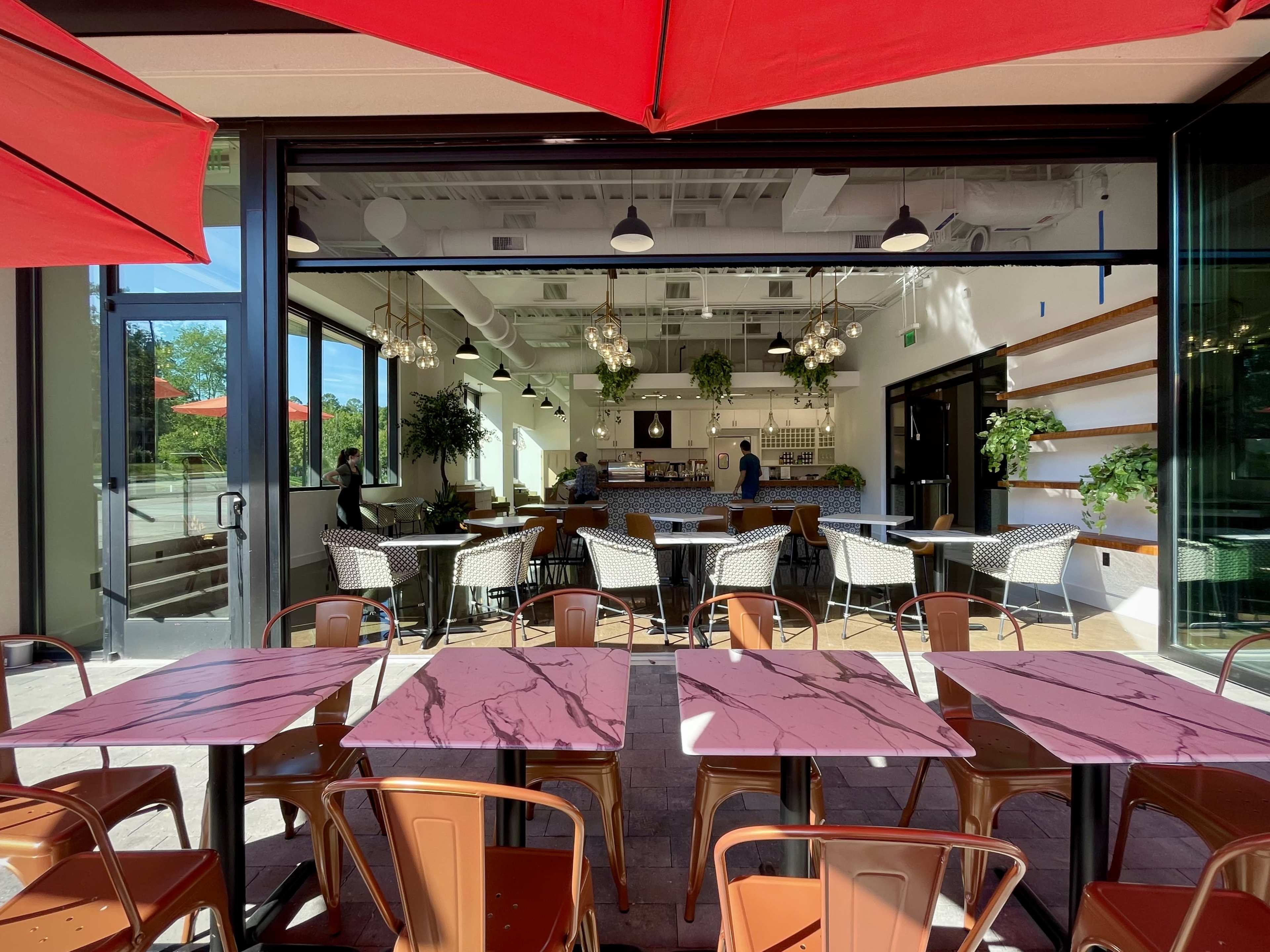 A modern café features outdoor tables with pink marble tops and red umbrellas, overlooking an open interior with a large seating area and greenery.