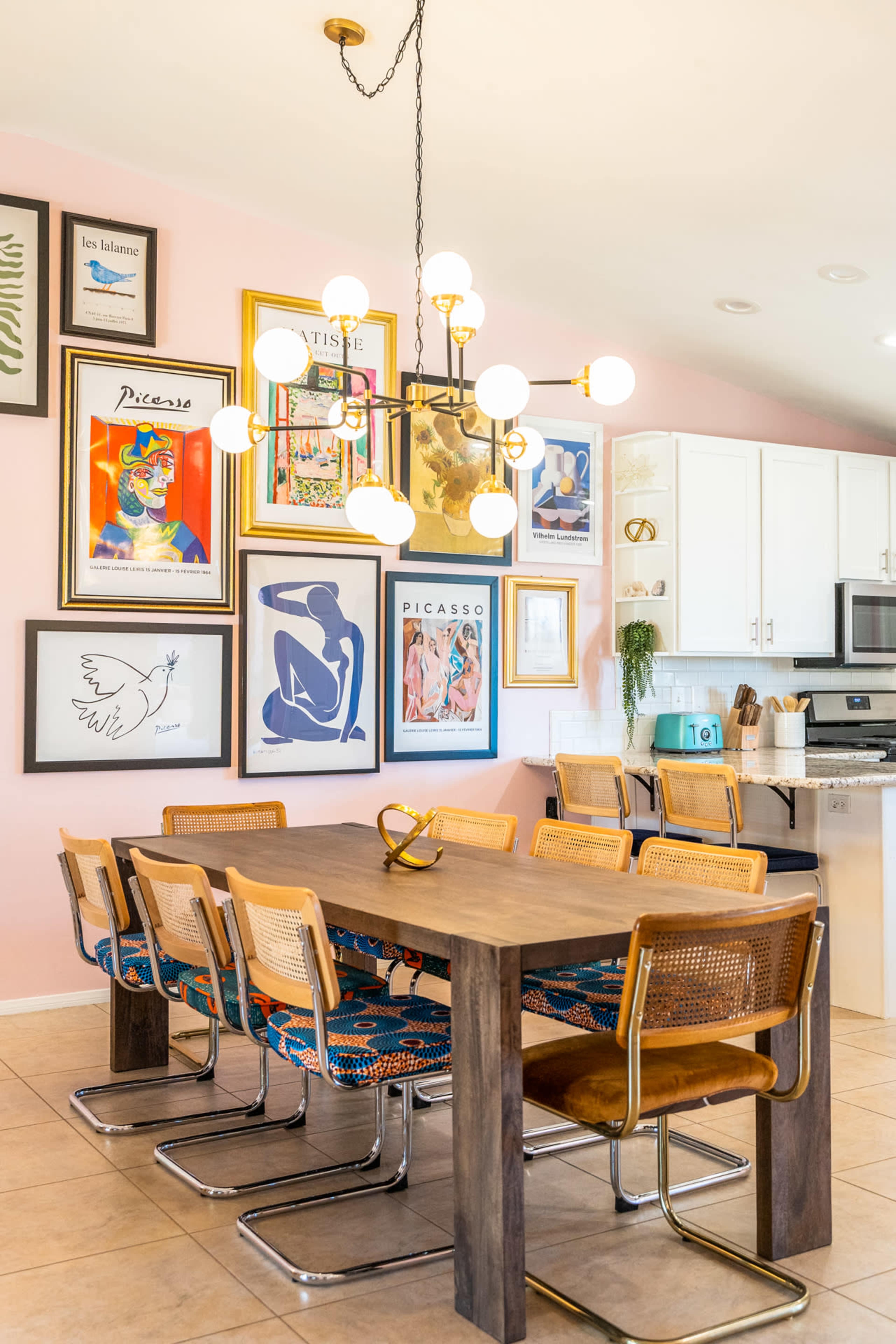A dining area features a wooden table surrounded by eight chairs, with a decorative chandelier above and an array of framed art on the wall.