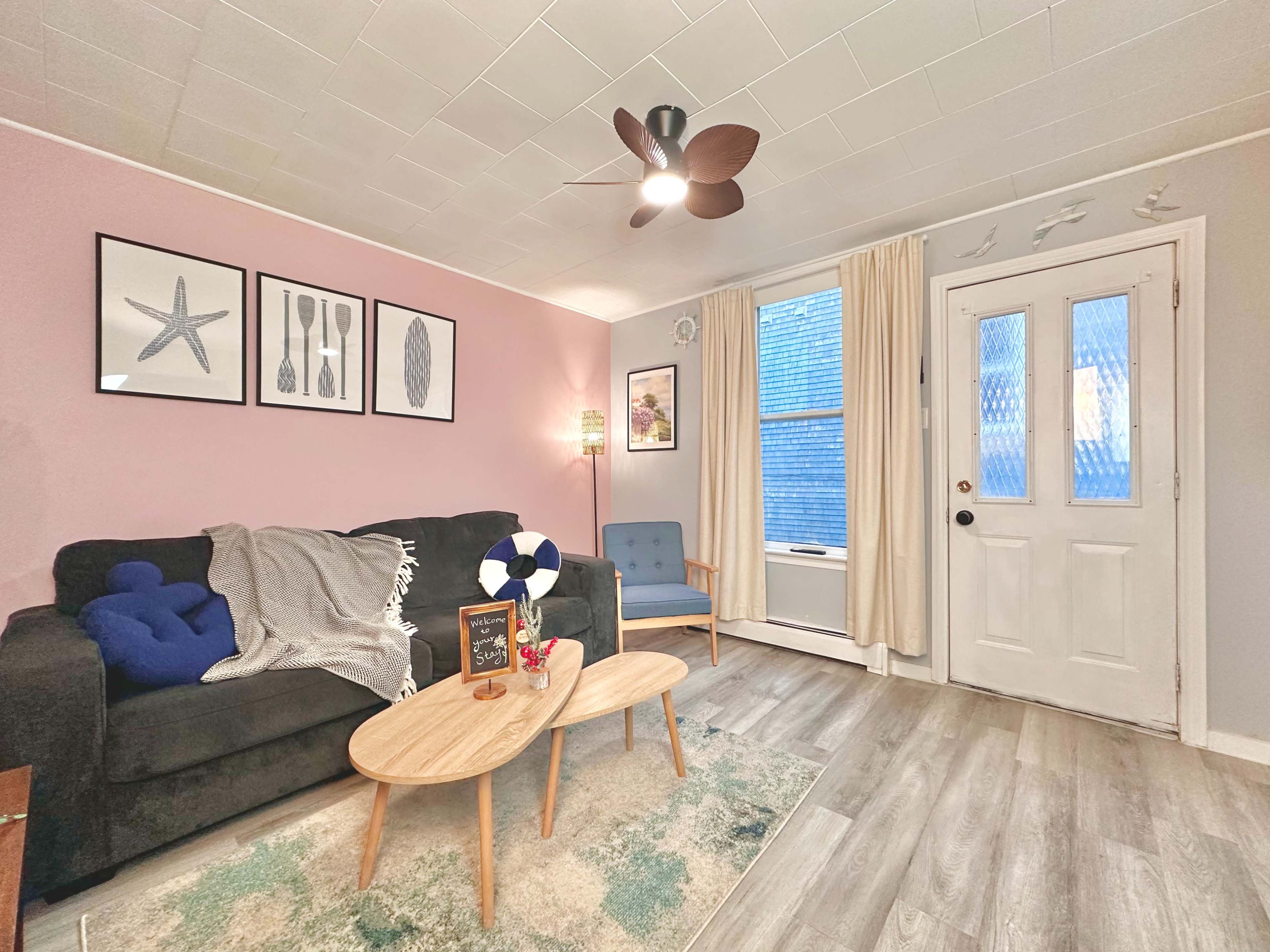 The image shows a cozy living room with a black couch, a blue armchair, a small wooden coffee table, and a door leading outside, accented by light pink walls and decorative wall art.