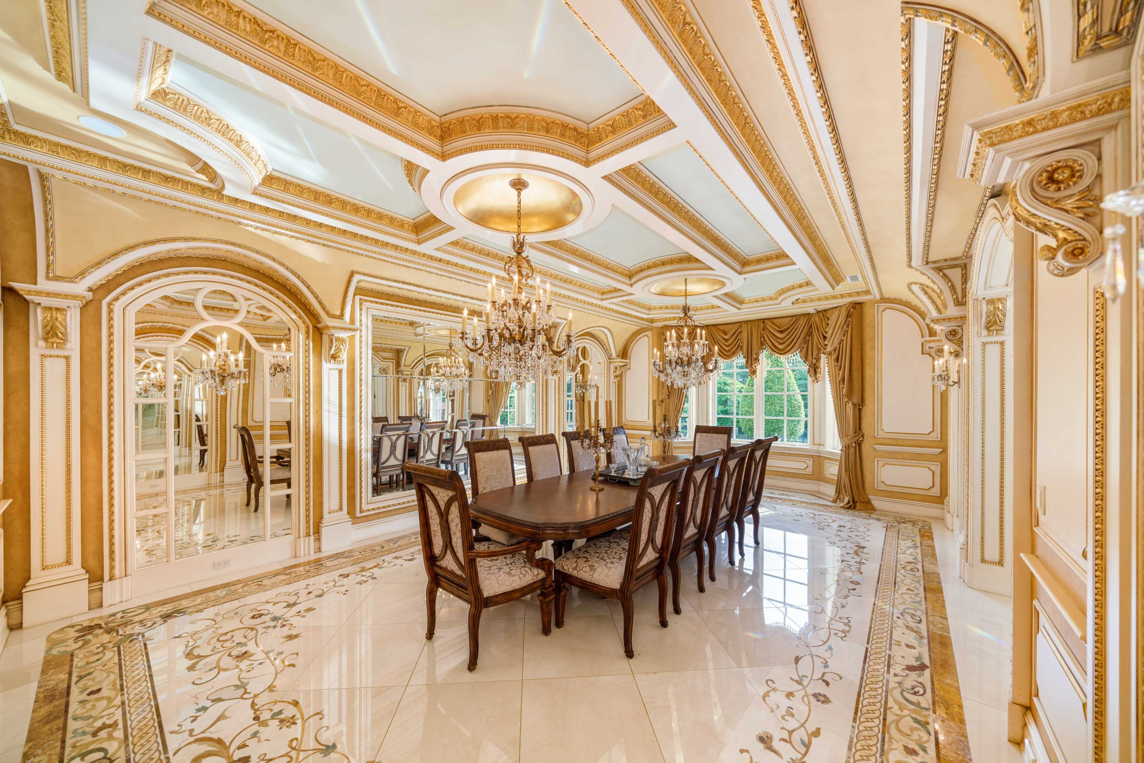 An ornate dining room featuring a large wooden table surrounded by upholstered chairs, with elaborate ceiling designs, golden accents, and large mirrors.