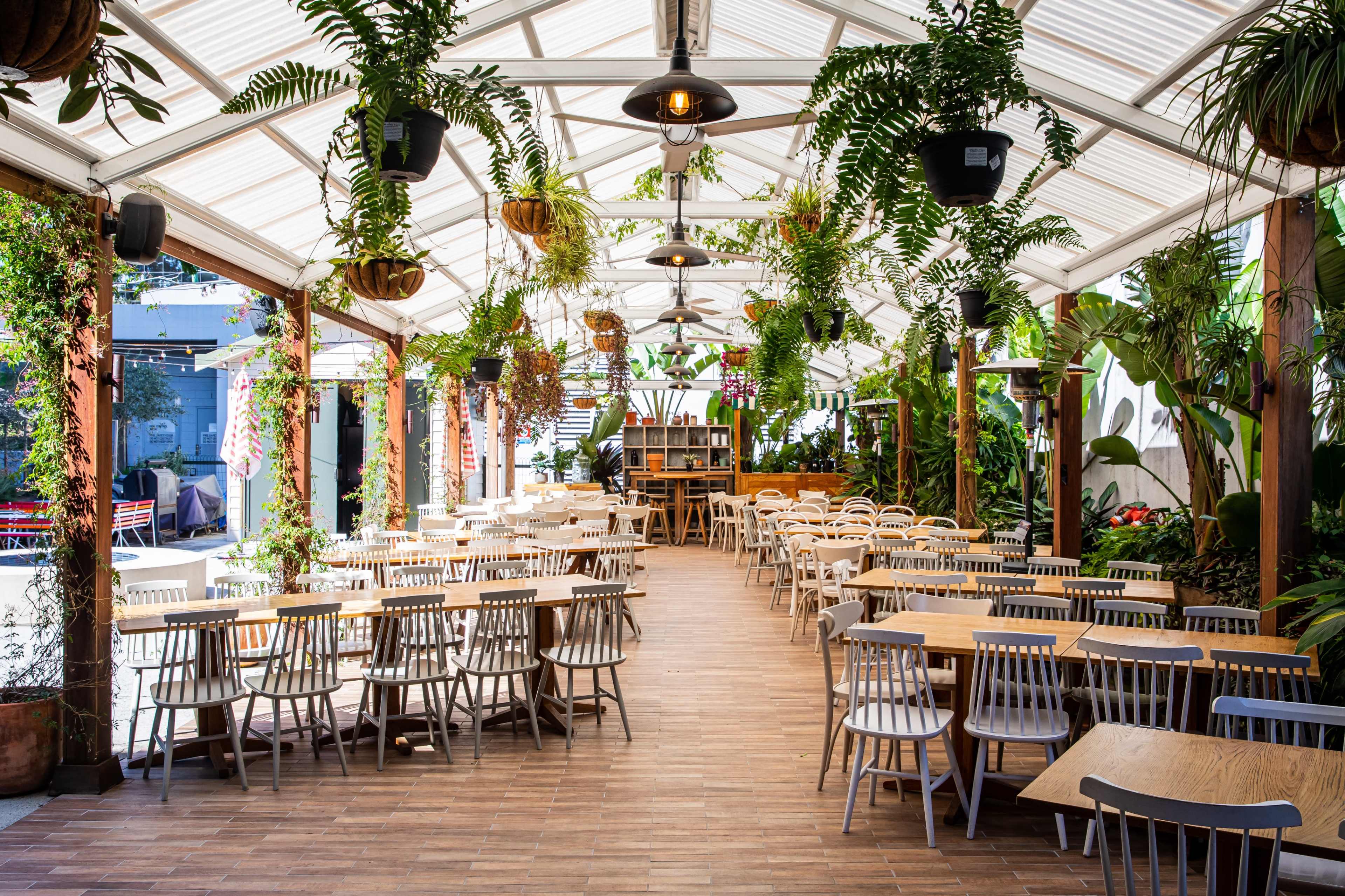 The image shows a spacious restaurant with a bright, covered dining area filled with wooden tables and chairs, surrounded by lush greenery and hanging plants.