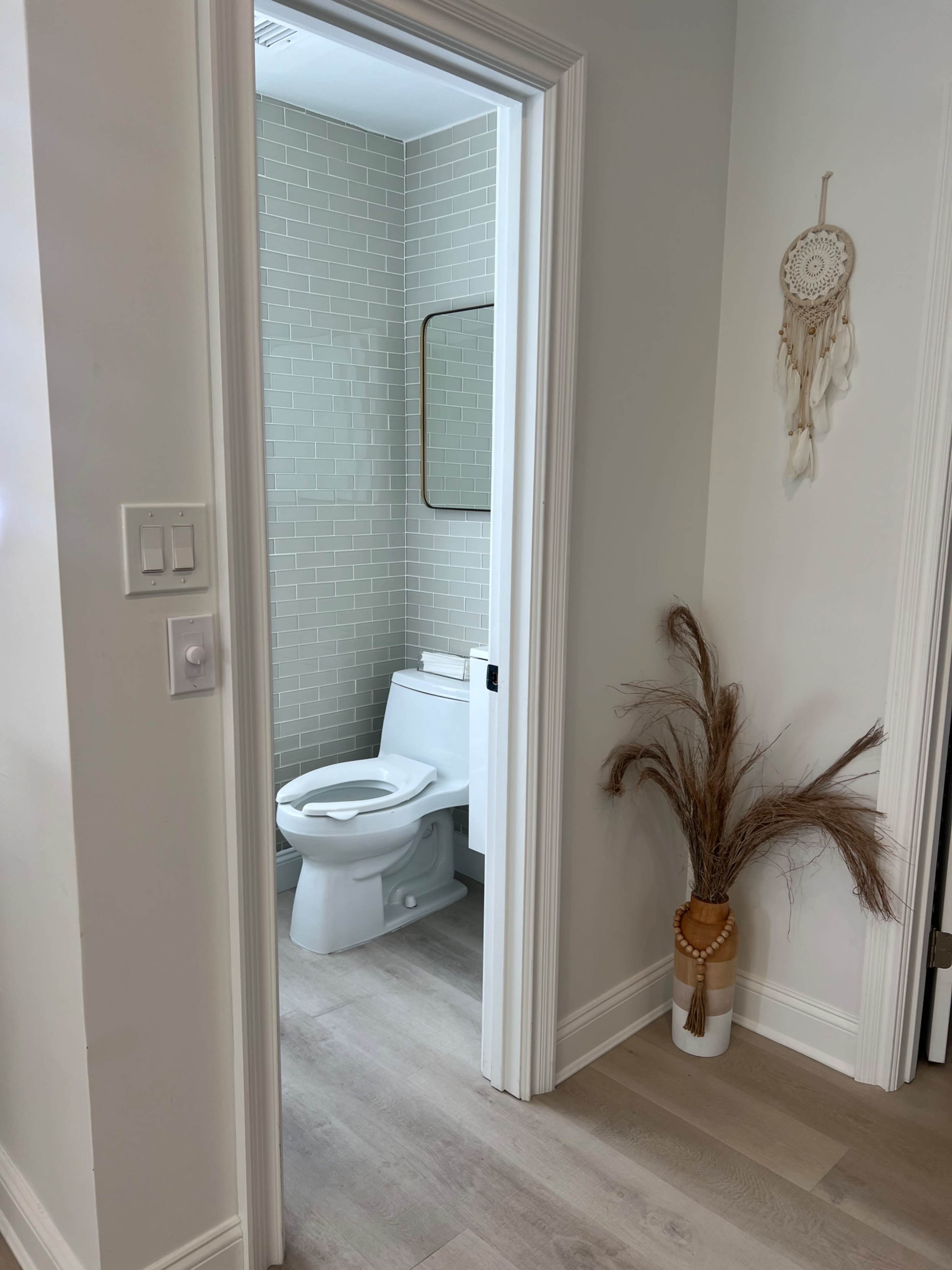 A clean bathroom is visible through an open doorway, featuring a toilet, a framed mirror, and a decorative plant in the foreground.
