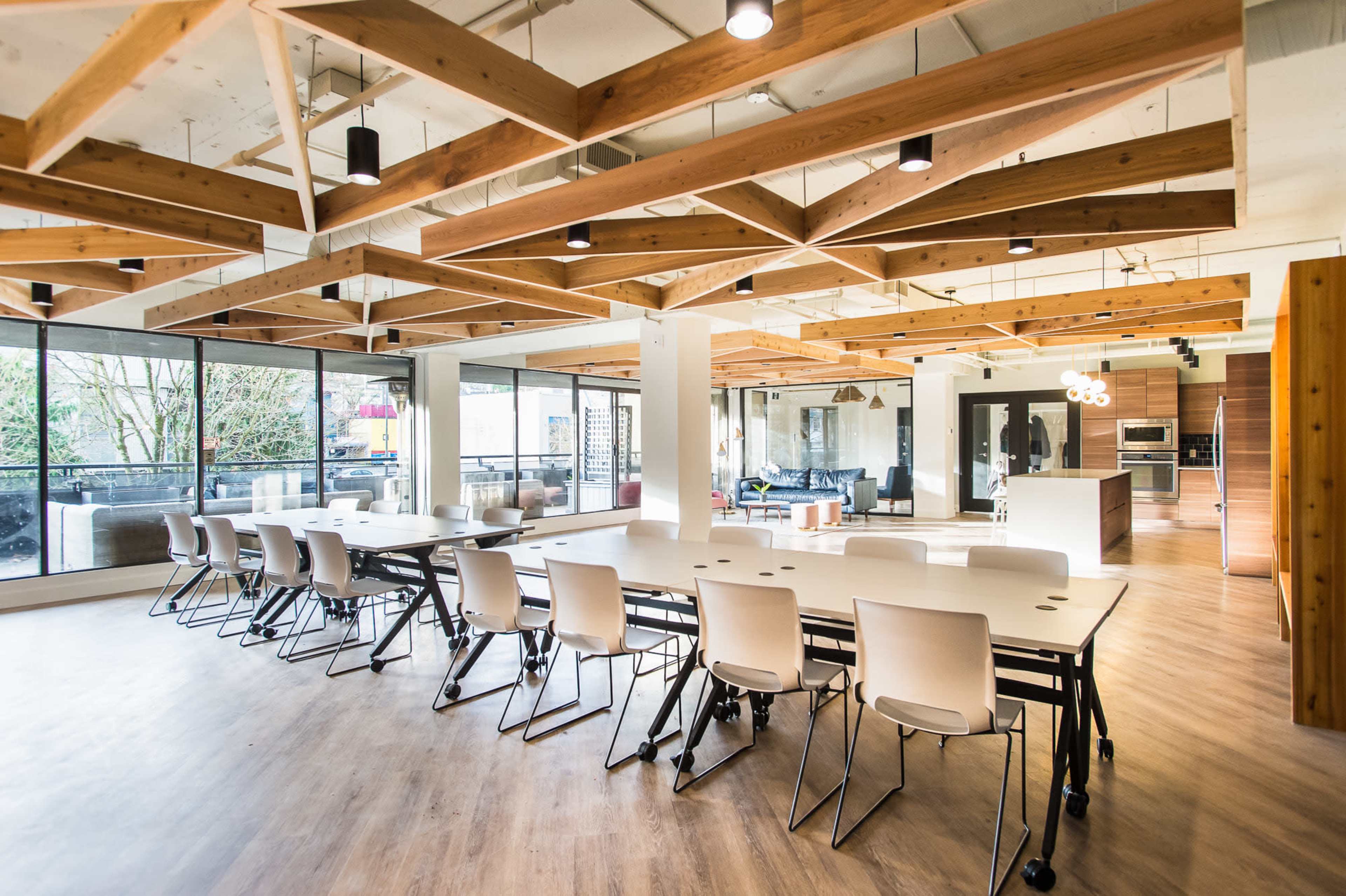 A spacious modern office with a large table, multiple chairs, and wooden beams in the ceiling, illuminated by natural light from large windows.